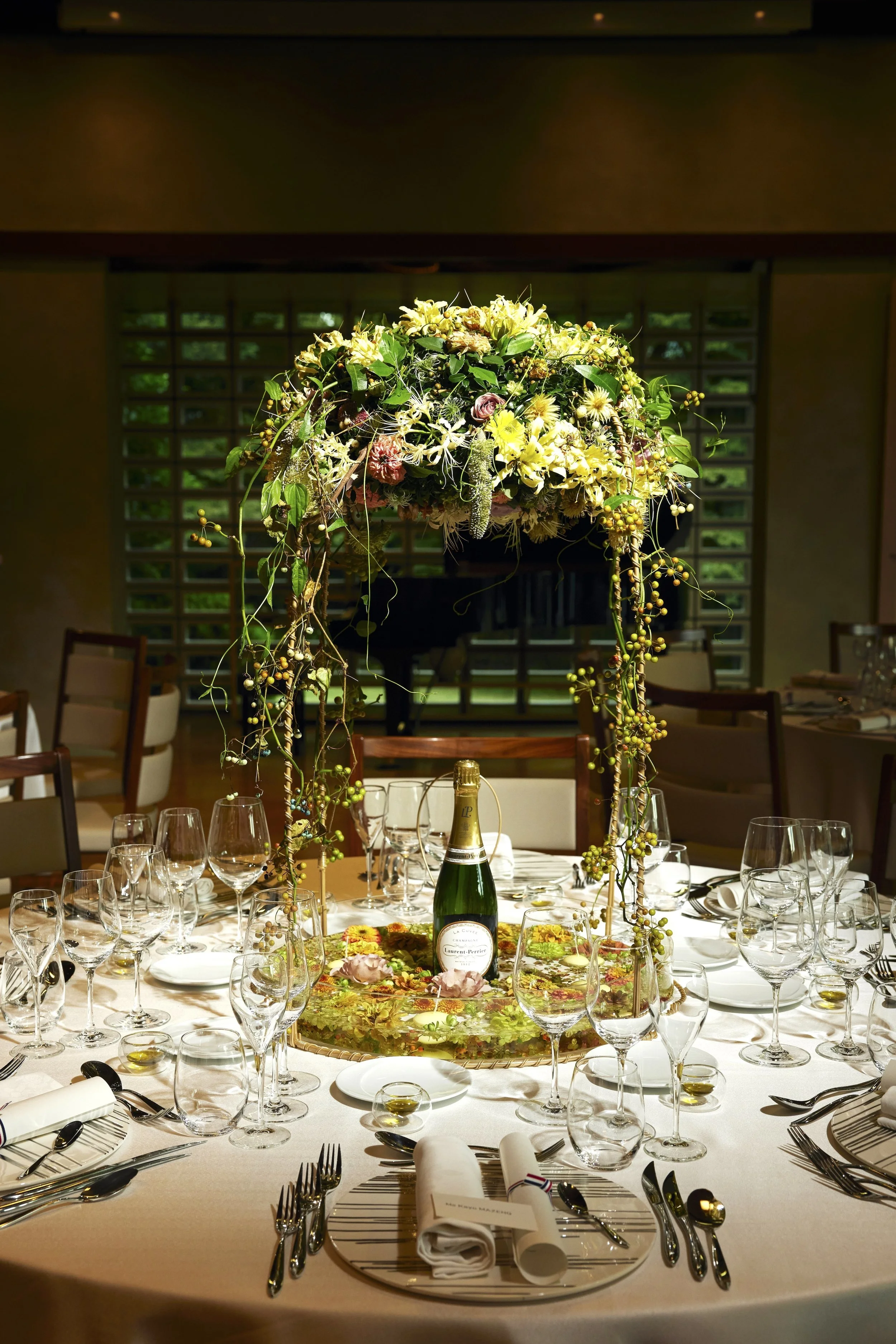 Round wedding table with a floral canopy centerpiece surrounding a bottle of champagne, combining luxury flowers and celebration in an intimate dining setting.