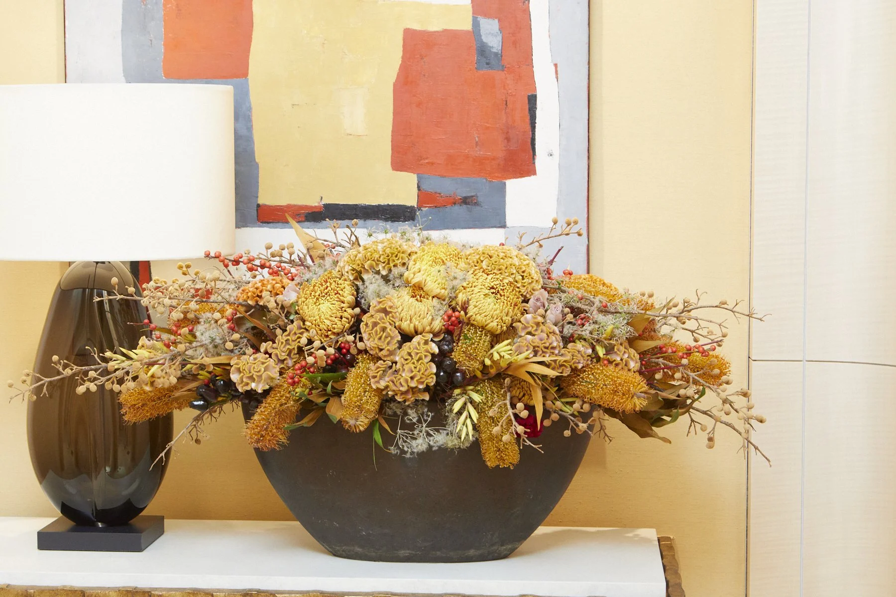 Low floral centerpiece with golden chrysanthemums, berries, and autumn textures in a black ceramic bowl, styled for a luxury hospitality interior.