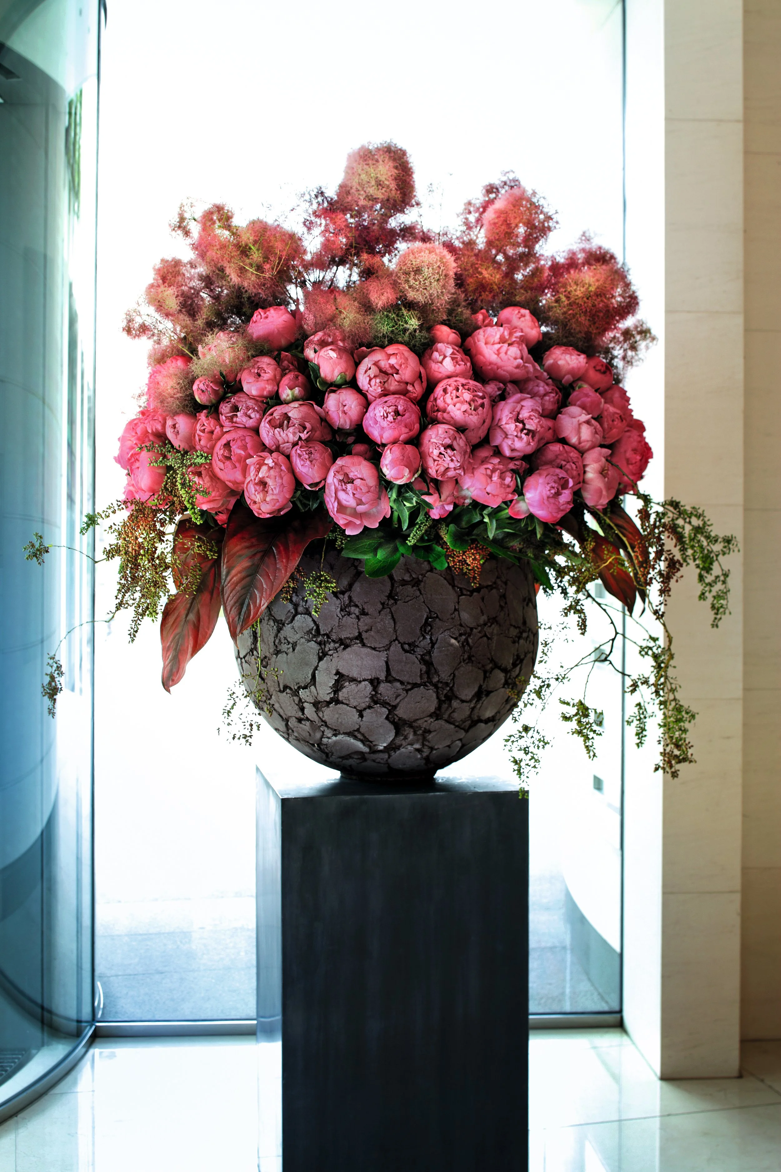 A large floral arrangement in a black crackled vase with pink peonies, greenery, and pink fluffy accents, displayed on a black pedestal in front of a glass door.