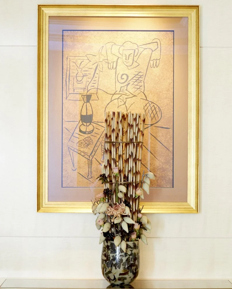 Architectural floral installation with vertical stems, berries, and soft blossoms arranged in a marble vase beneath a framed artwork in a hotel suite.