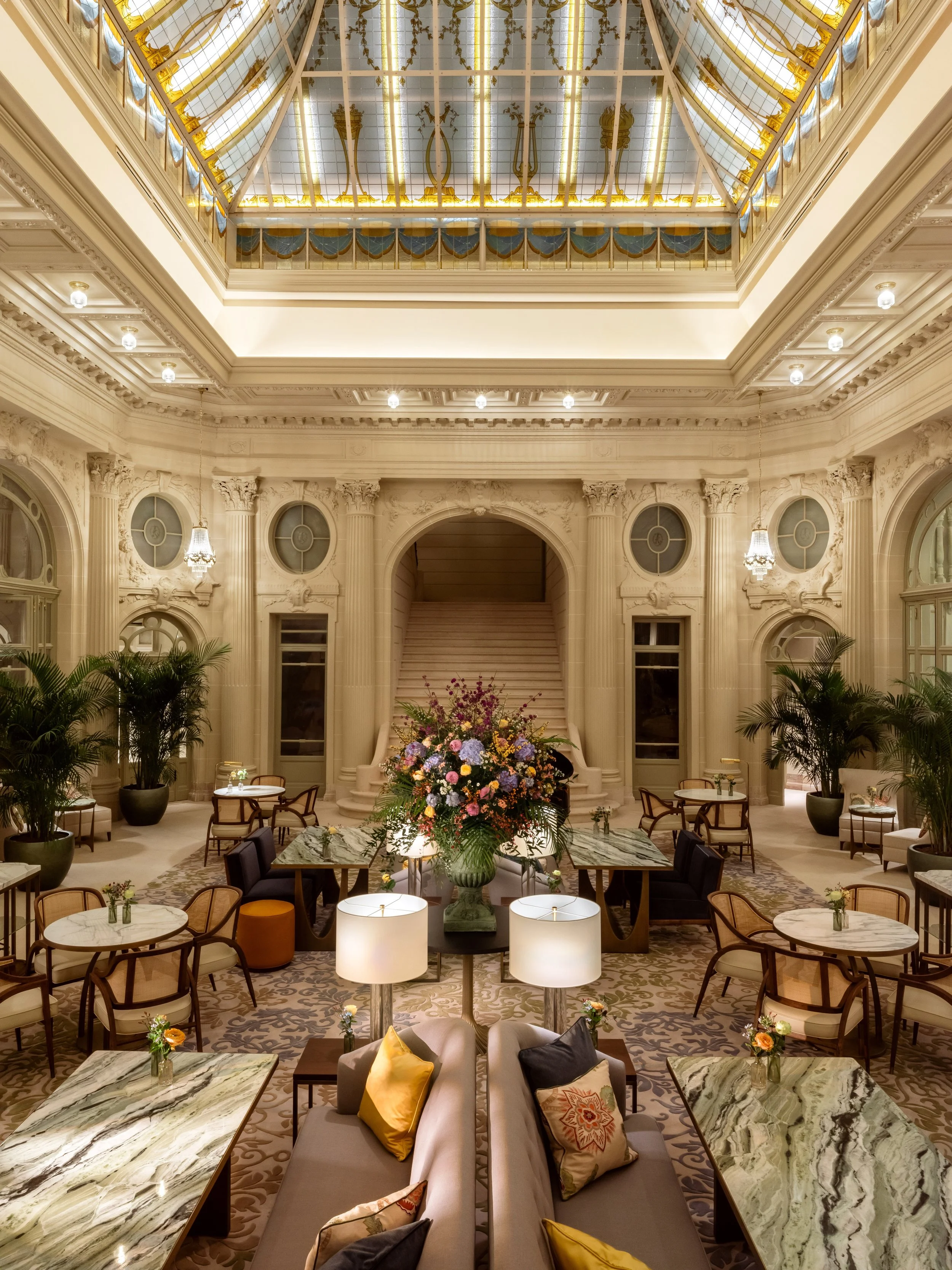 Grand floral centerpiece in a hotel atrium lounge, with tall vase of mixed seasonal blooms under a stained-glass dome ceiling.