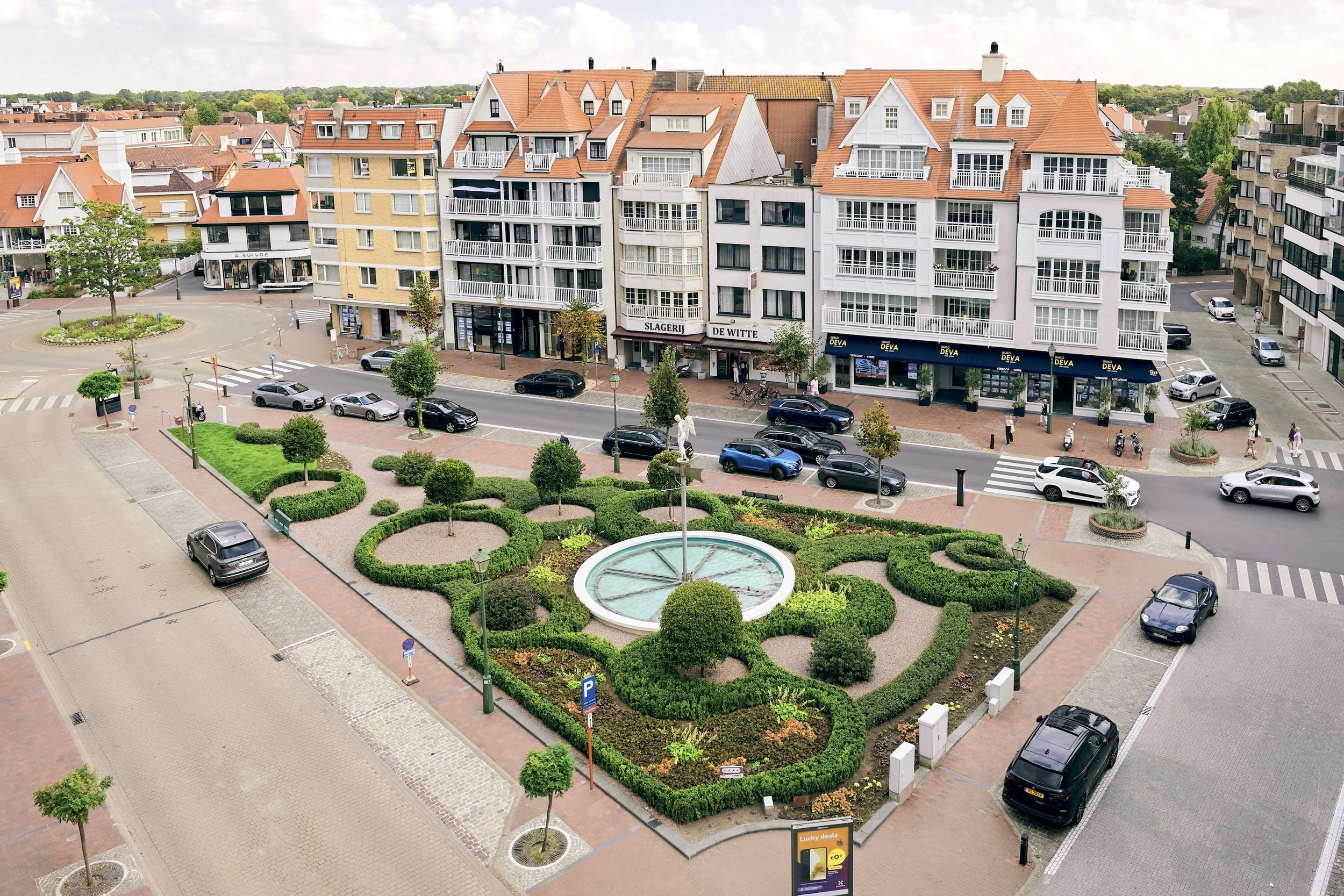 Town square with circular fountain and manicured topiary garden surrounded by residential and commercial buildings.