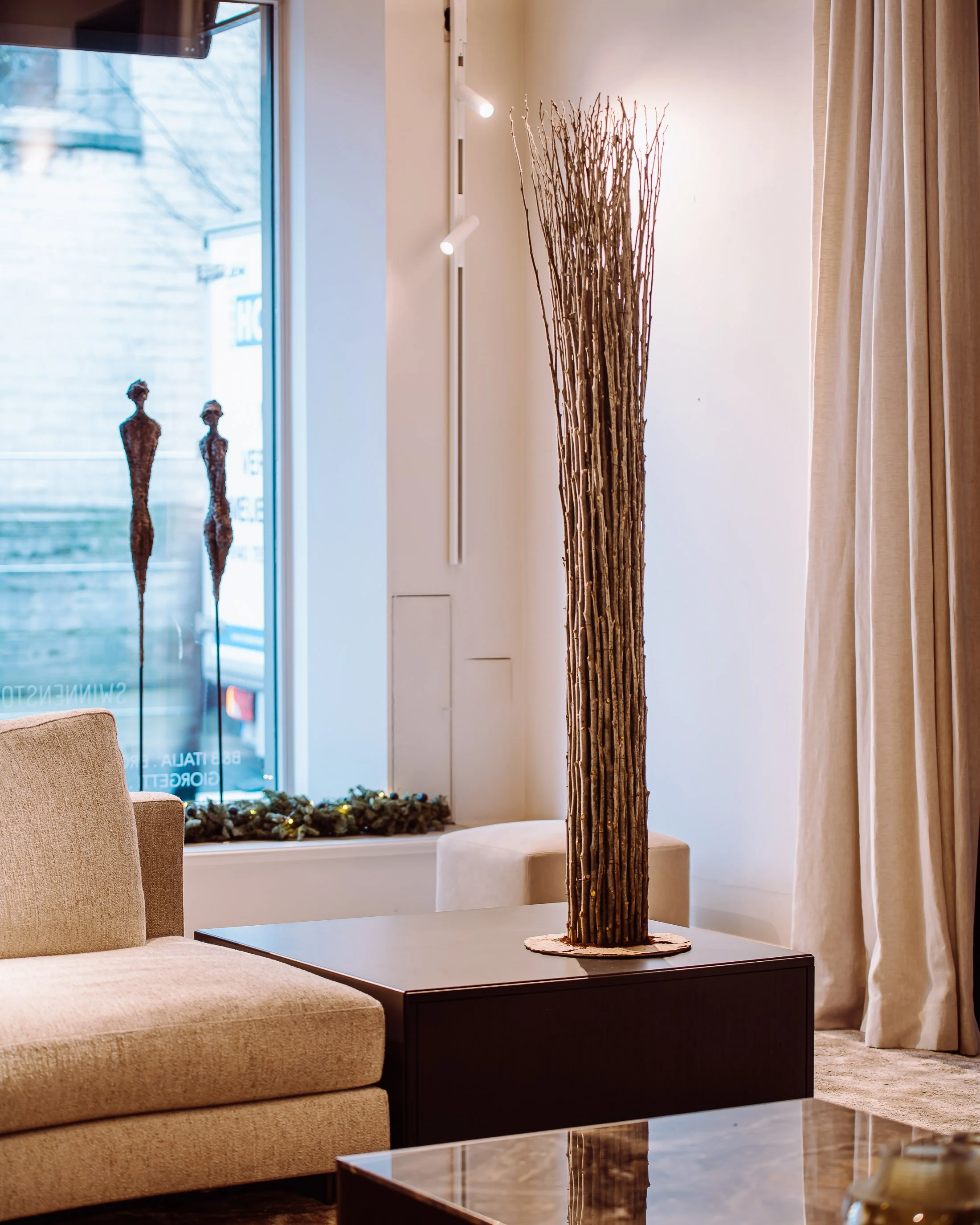 Interior of a room with beige sofas, a dark coffee table, a tall decorative dried branch arrangement on a black stand, and a window with curtains. Outside the window, two abstract sculptures are visible.