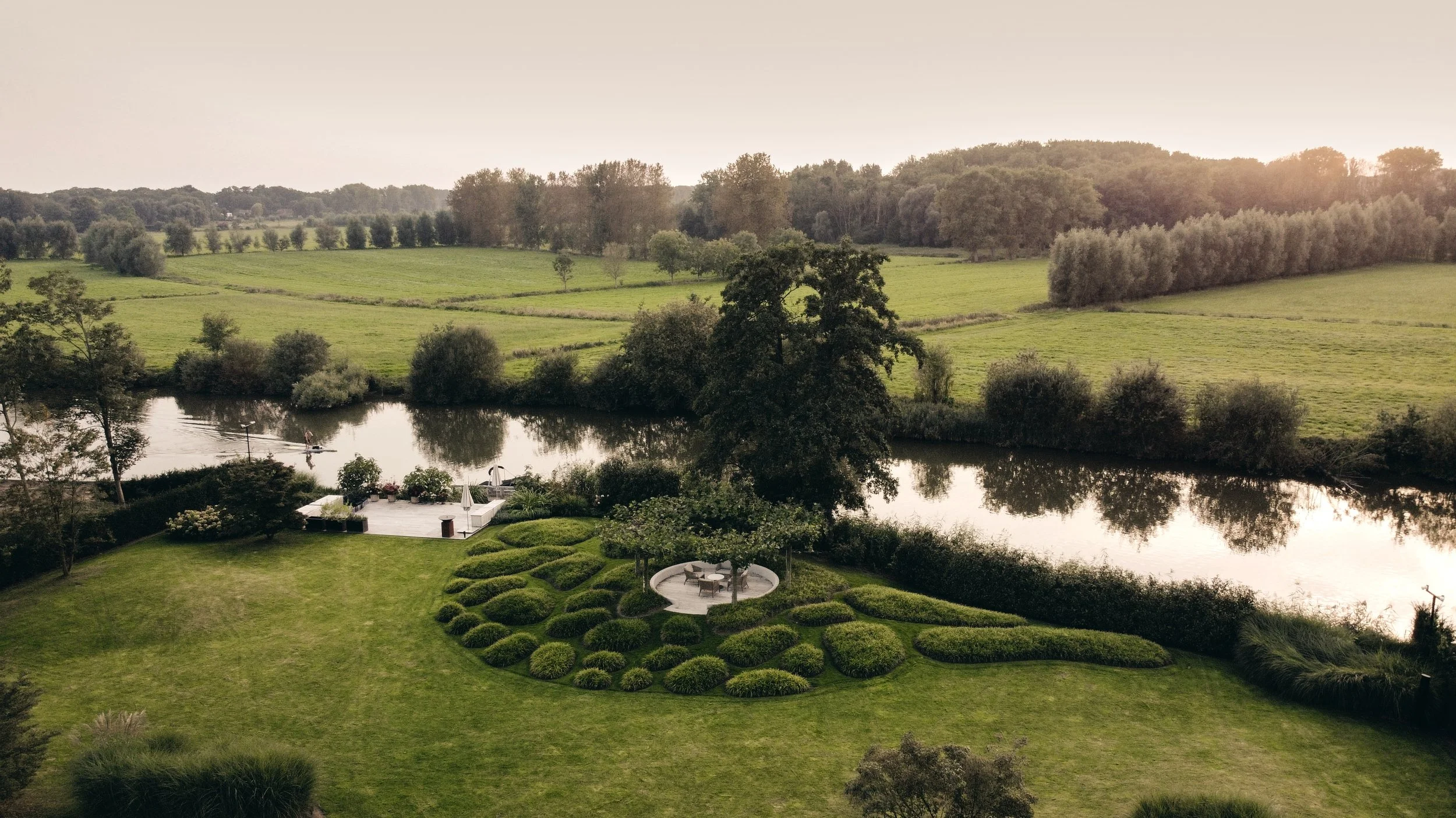Riverside garden with organic topiary design – Sculpted lawn mounds and shaded seating pavilion along a tranquil riverbank with wide countryside views.