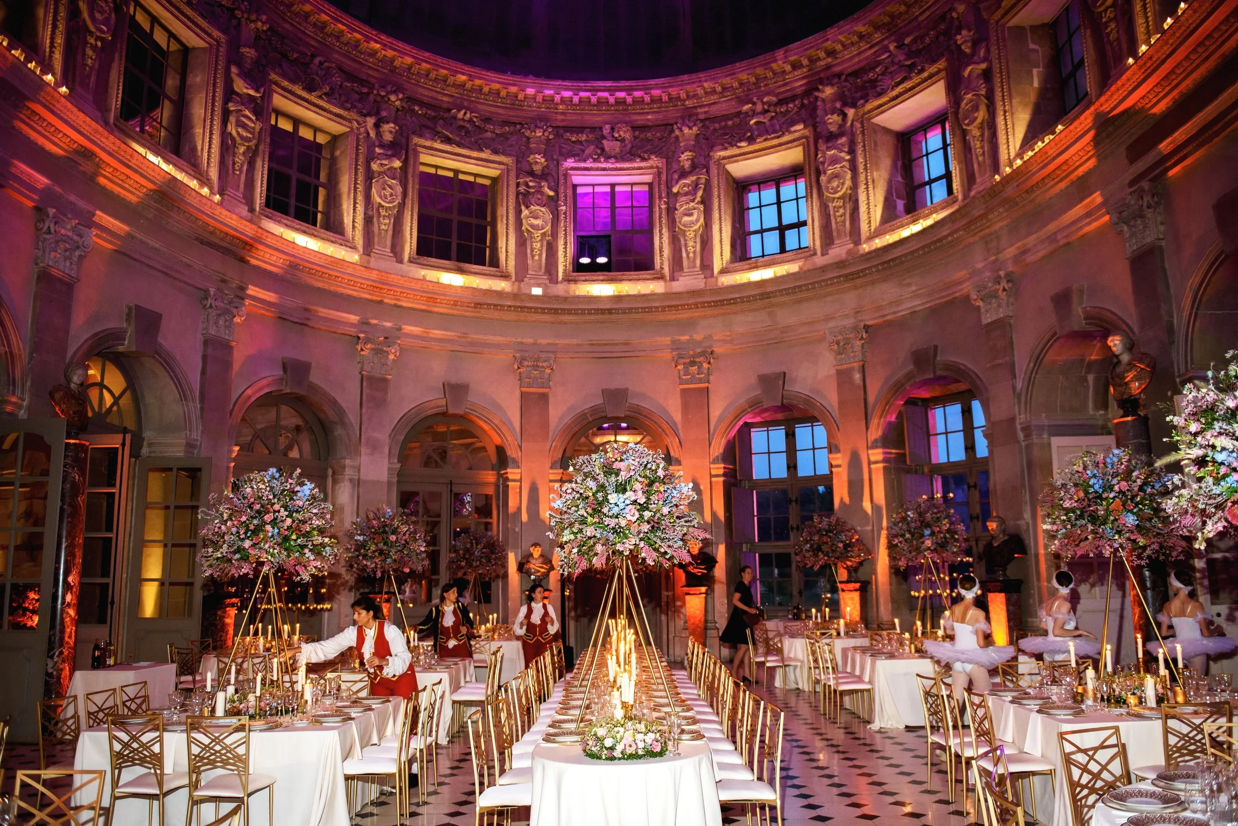 Grand wedding dinner in a historic palace ballroom, with towering pastel floral centerpieces, candlelit tables, and opulent golden architecture.