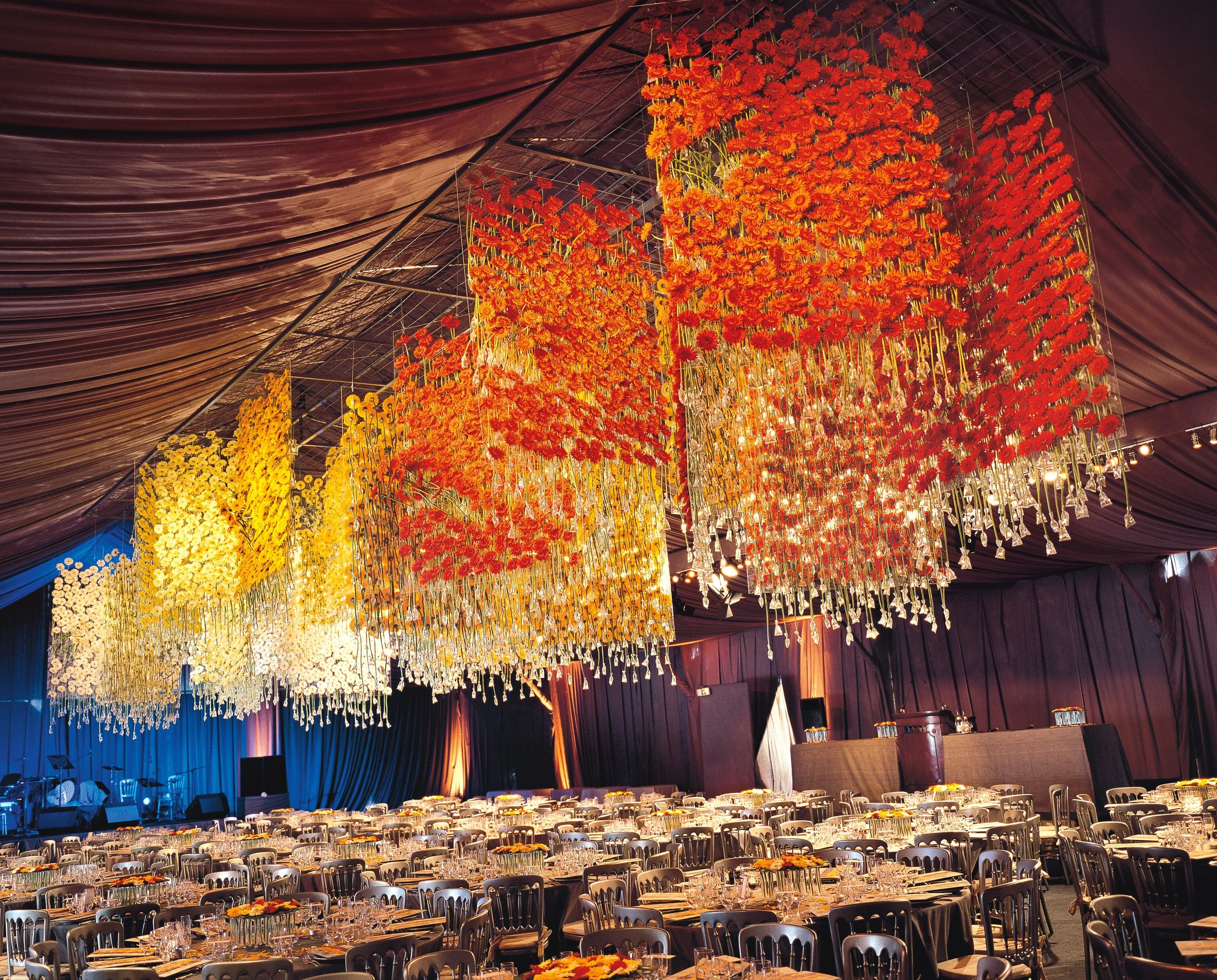 Corporate gala with striking ceiling installation of floating orange and yellow flower panels, illuminated to create a vibrant floral canopy by Daniel Ost