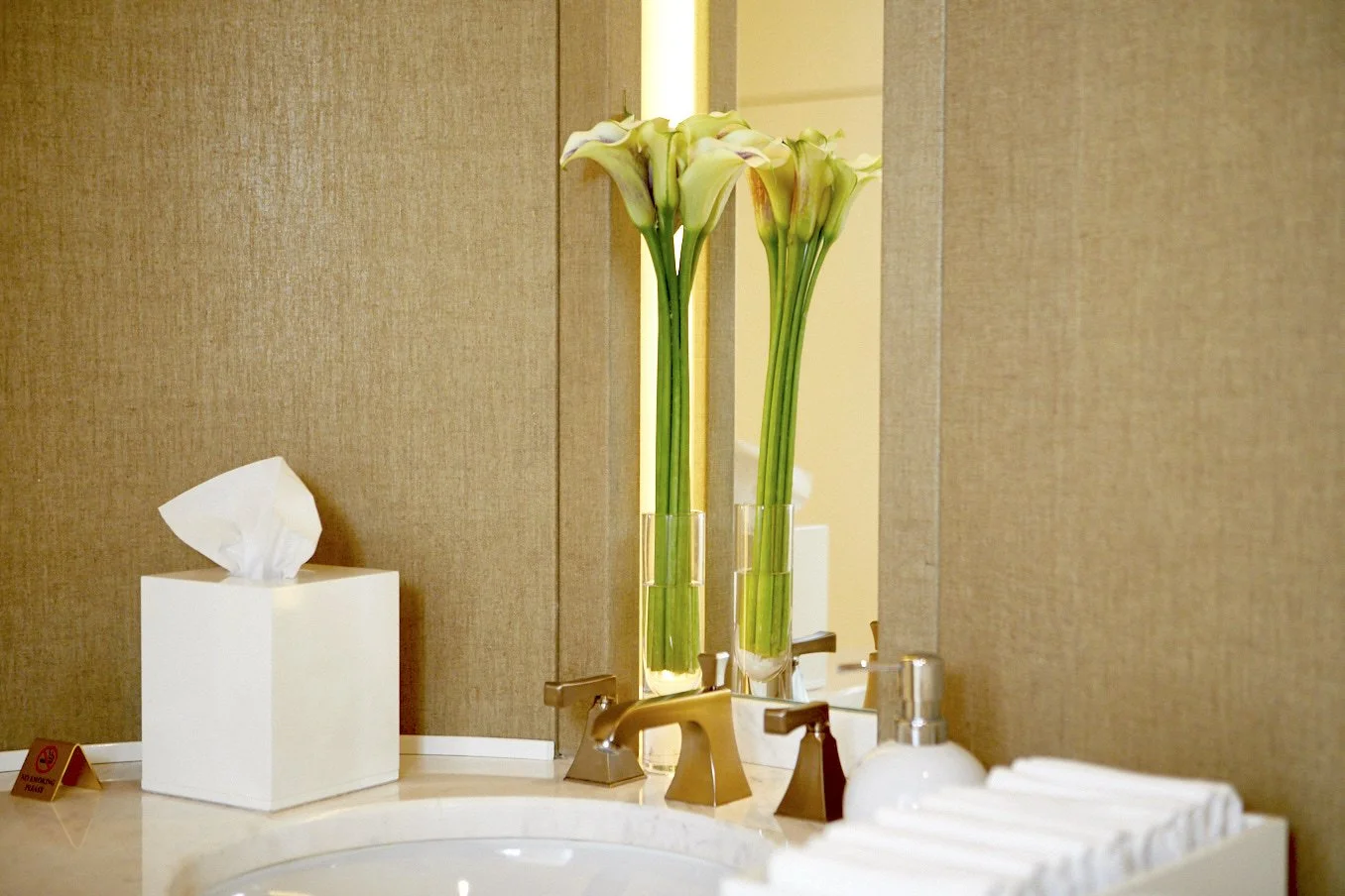 Elegant hospitality washroom floral detail with tall white calla lilies in slim glass vases placed on a marble sink counter.