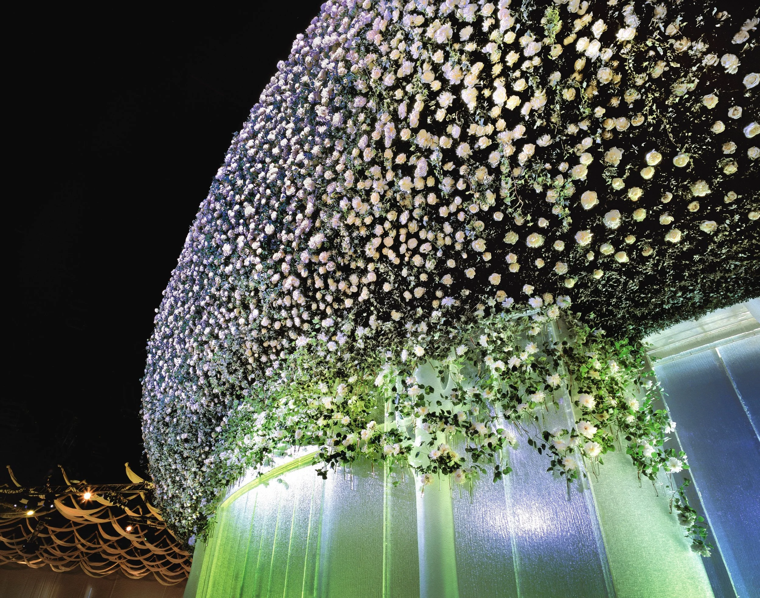 Luxury wedding stage design with thousands of cascading white roses forming a circular floral canopy above shimmering drapery and lights — a sculptural floral installation by Daniel Ost.