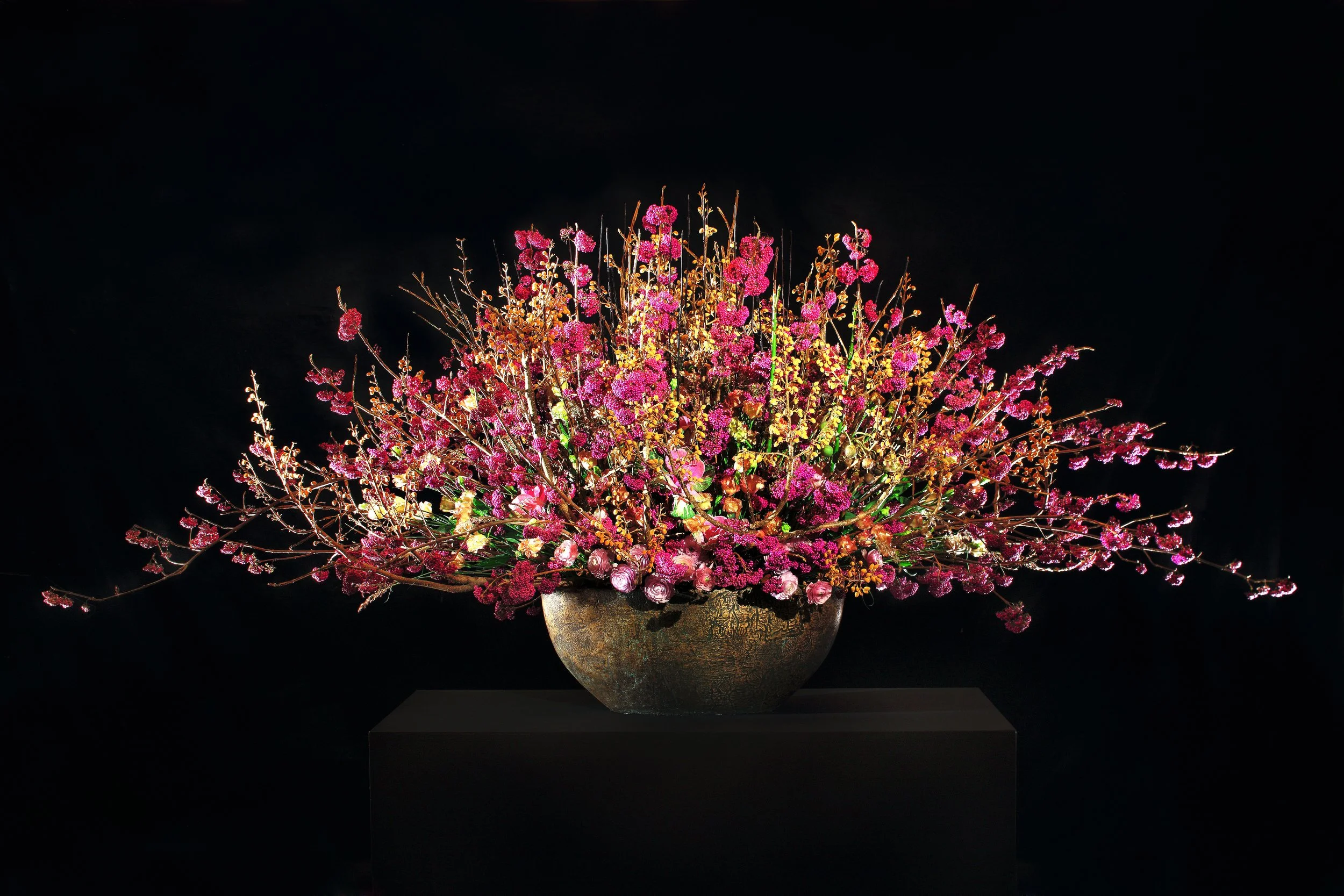 A large, elaborate floral arrangement with pink flowers and golden branches in a textured vase against a dark background.