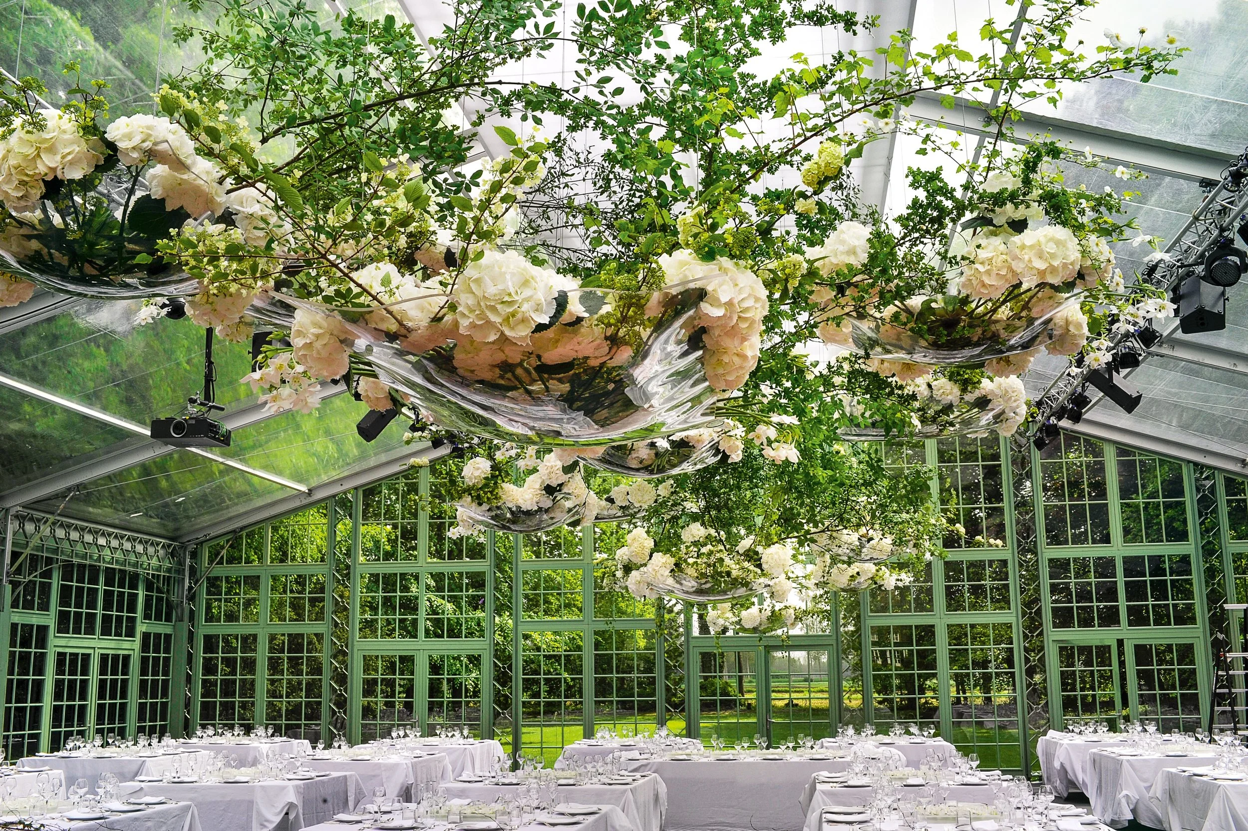 Transparent greenhouse venue with suspended sculptural floral clouds of hydrangeas and greenery, floating above elegant white wedding banquet tables