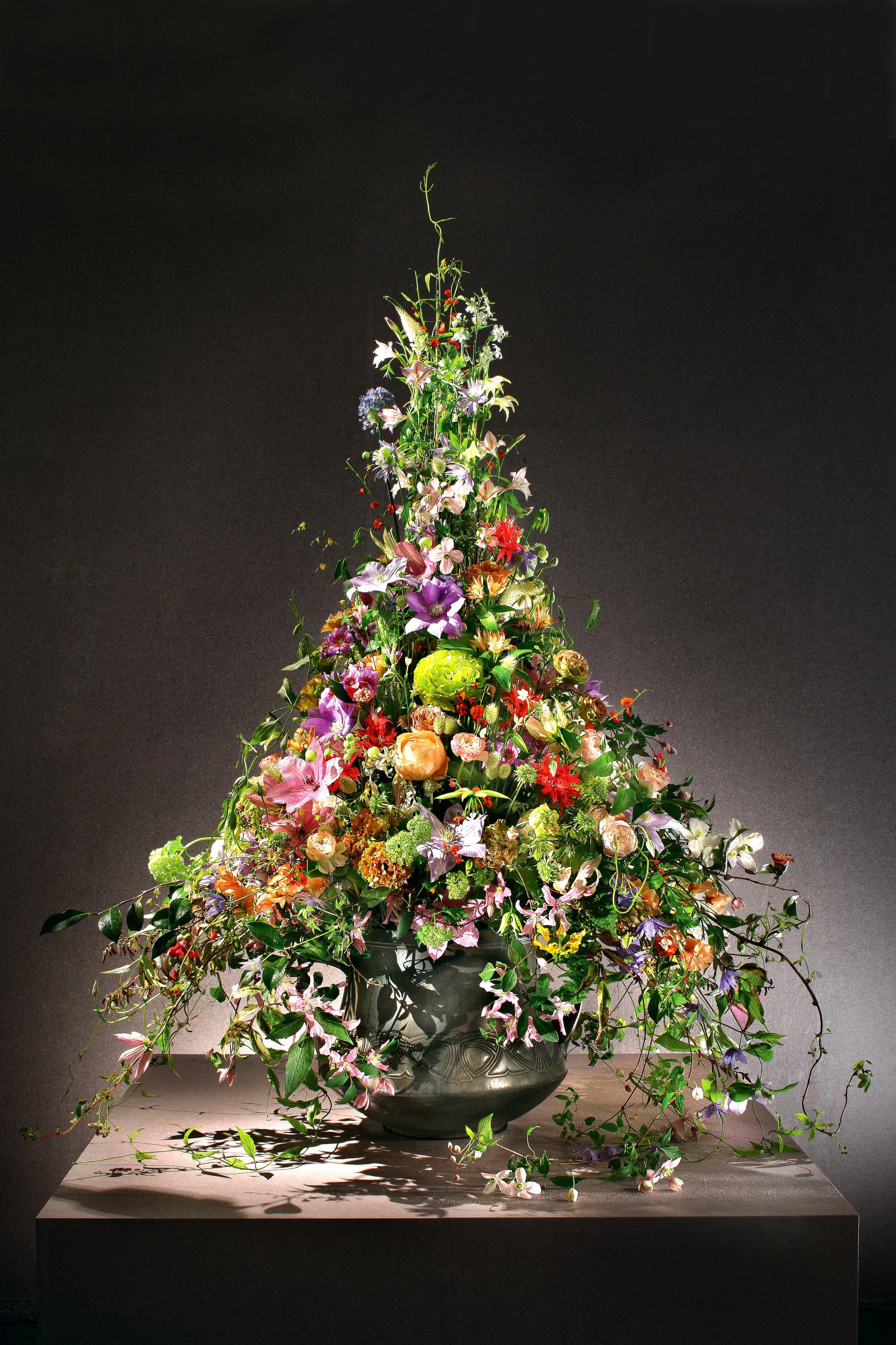 A large, elaborate flower arrangement in a dark decorative vase, featuring various colorful flowers and greenery, displayed on a pedestal against a dark background.