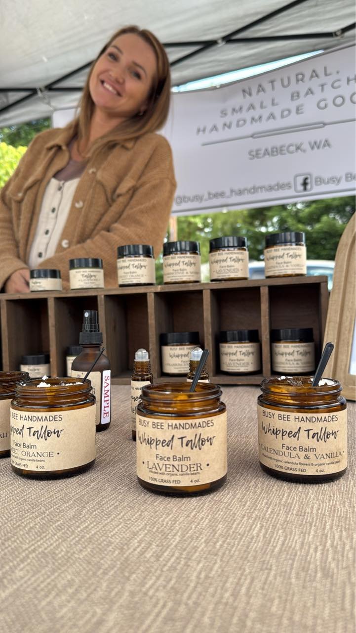 Woman standing behind a table displaying handmade whipped shea butter face balms at an outdoor market booth.