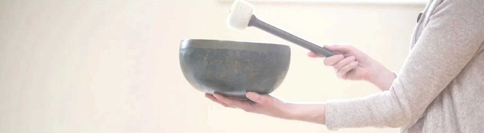 Person holding a metal singing bowl with one hand and striking it with a mallet in the other hand.