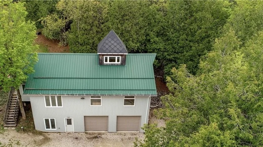 A white garage building with a green metal roof and a small dormer window, surrounded by dense green trees.