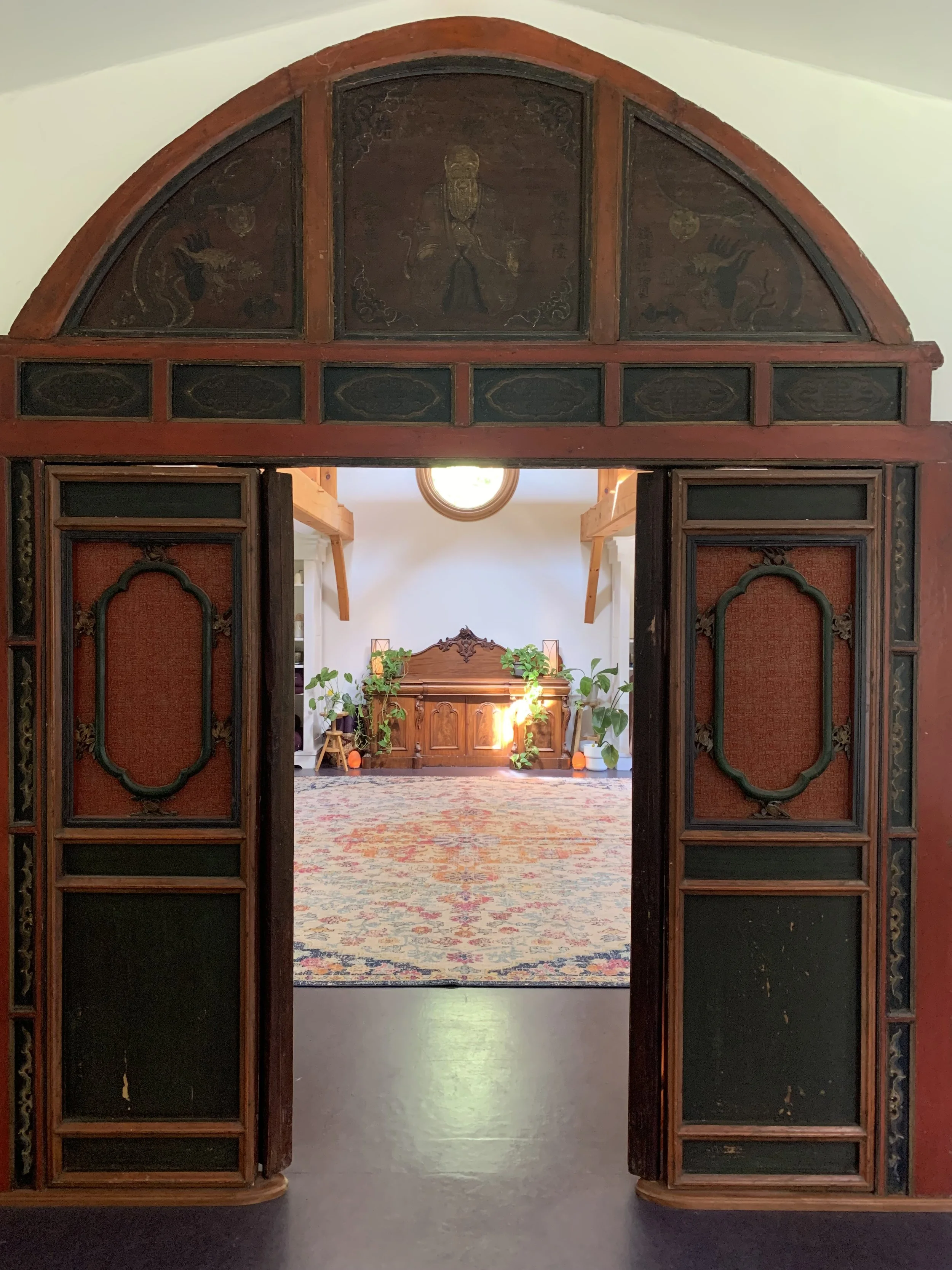 View through a vintage wooden doorway into a room with a wooden headboard, large potted plants, and a colorful rug on the floor.