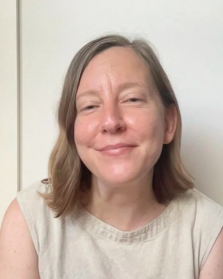 A woman with shoulder-length light brown hair smiling at the camera, wearing a beige sleeveless top, against a plain light-colored wall background.