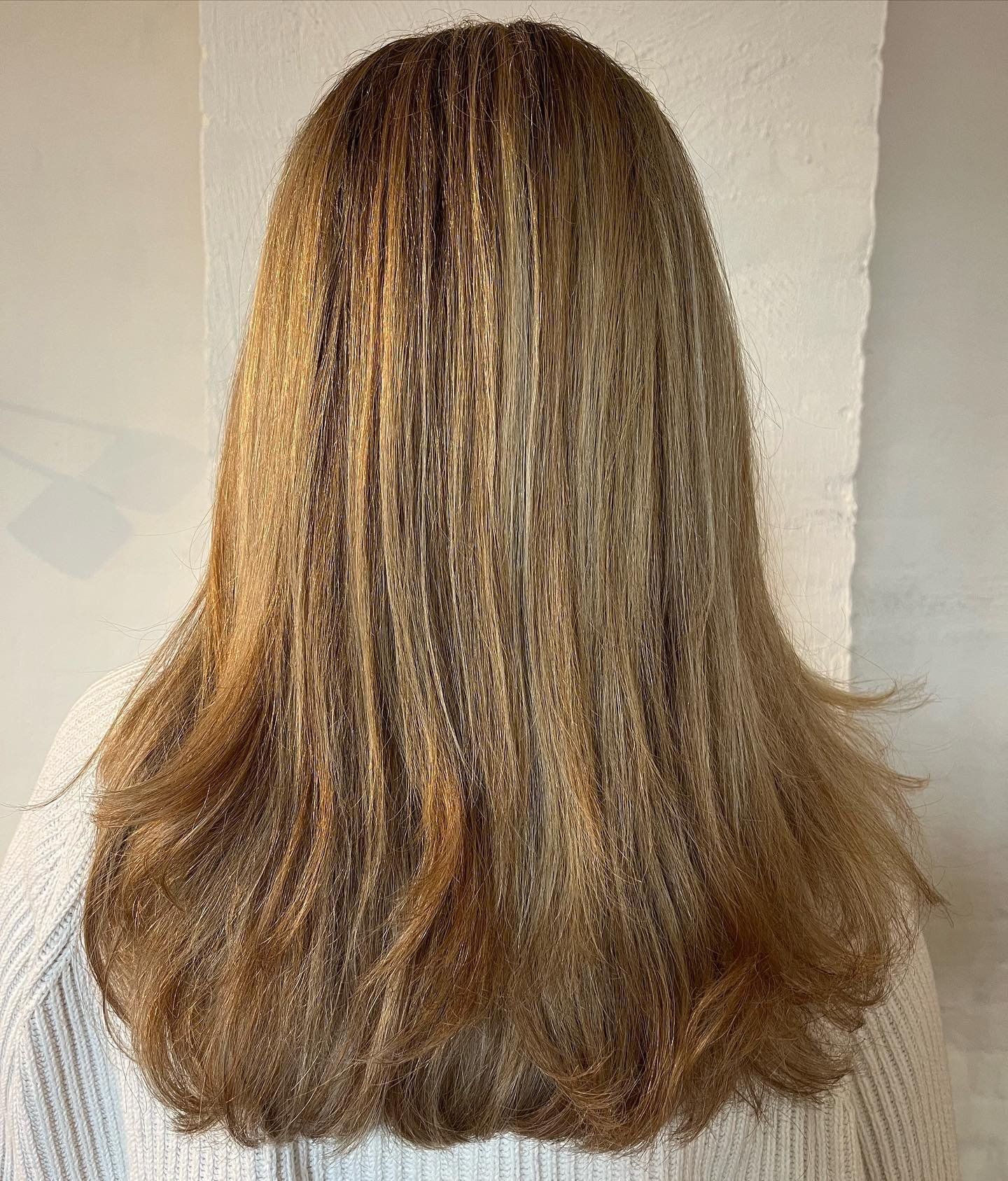 Back view of a woman with shoulder-length, straight, light brown hair with subtle highlights, wearing a white sweater, standing against a light-colored wall.