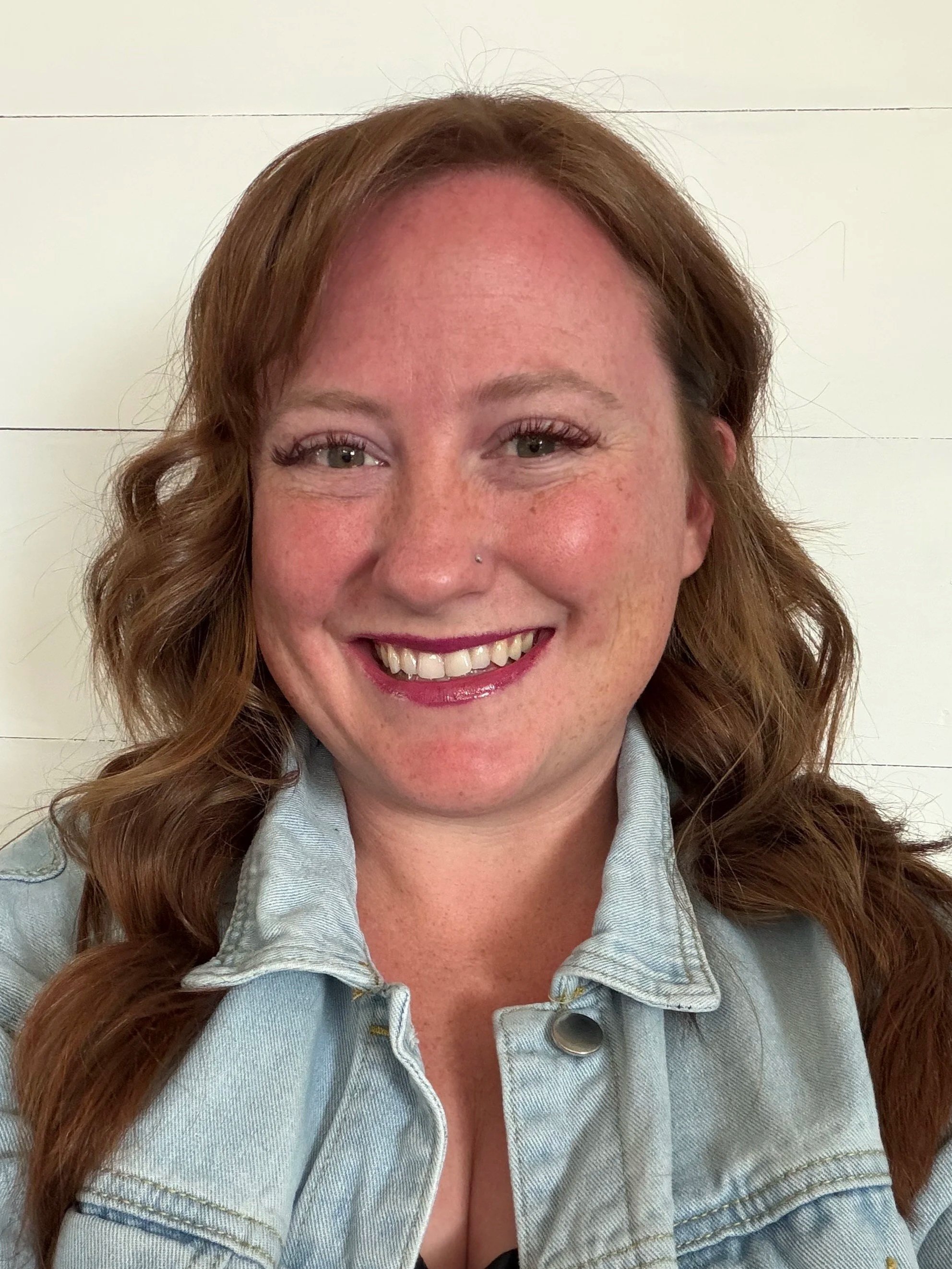 A woman with red hair, freckles, and a nose piercing smiling at the camera, wearing a light denim jacket against a white background.
