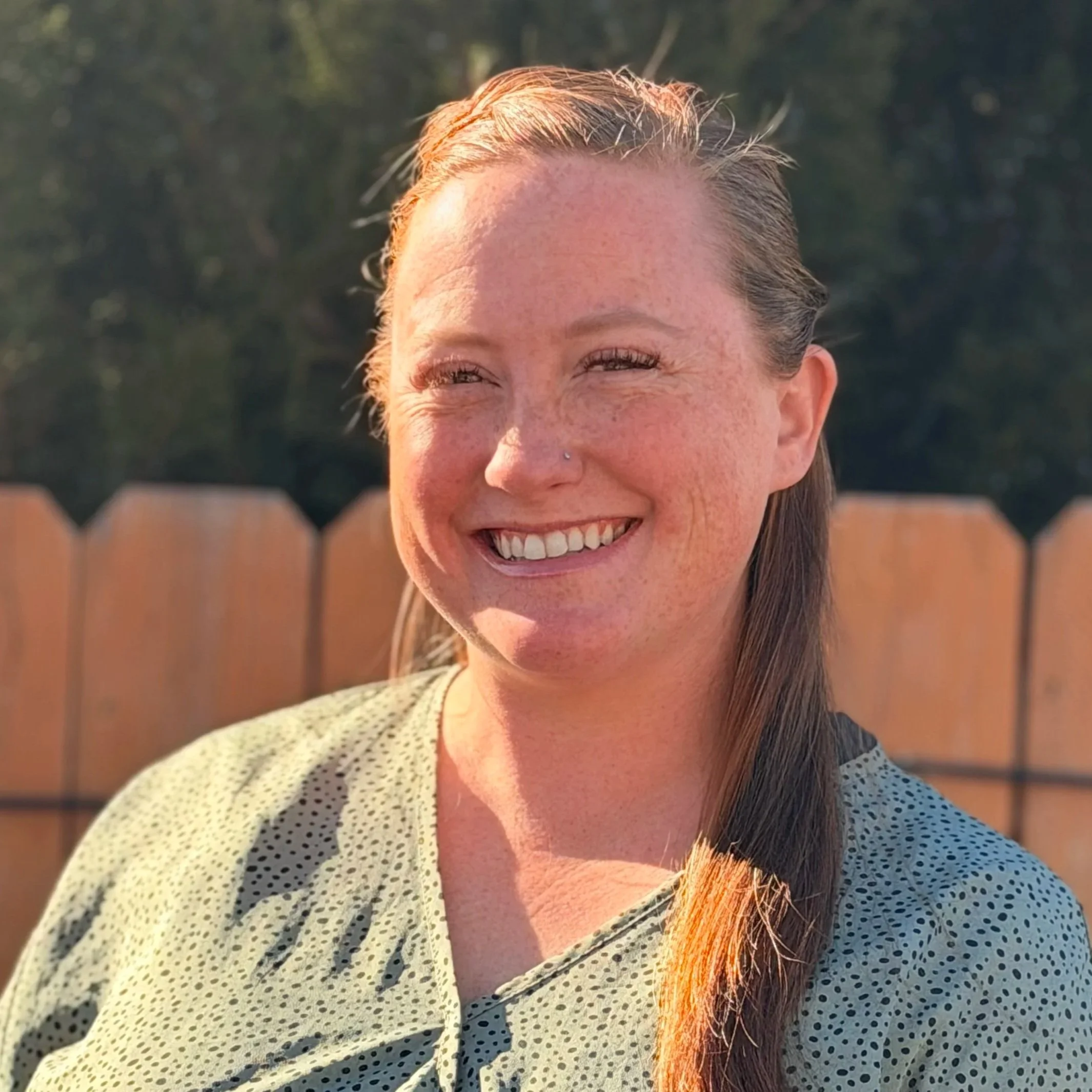 A young woman with long brown hair tied to one side, smiling outside in front of a wooden fence and greenery, in sunlight.
