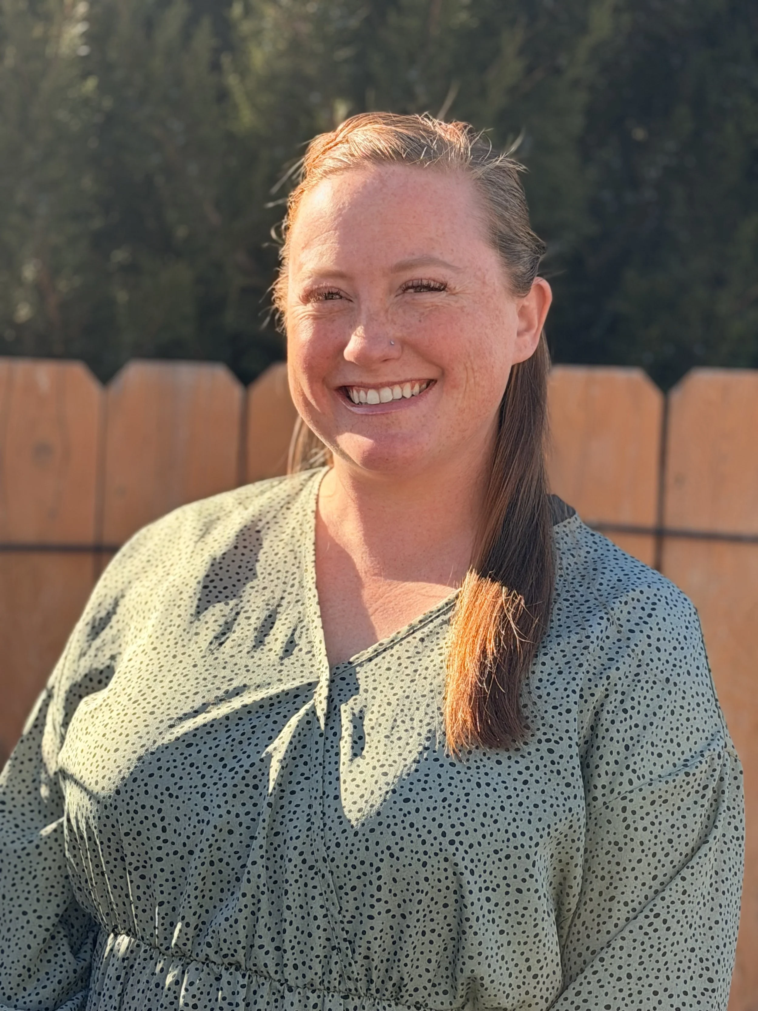 A smiling woman with light skin, freckles, and a nose piercing standing outdoors in front of a wooden fence and trees, wearing a light green dress with black polka dots and clasped at the waist.