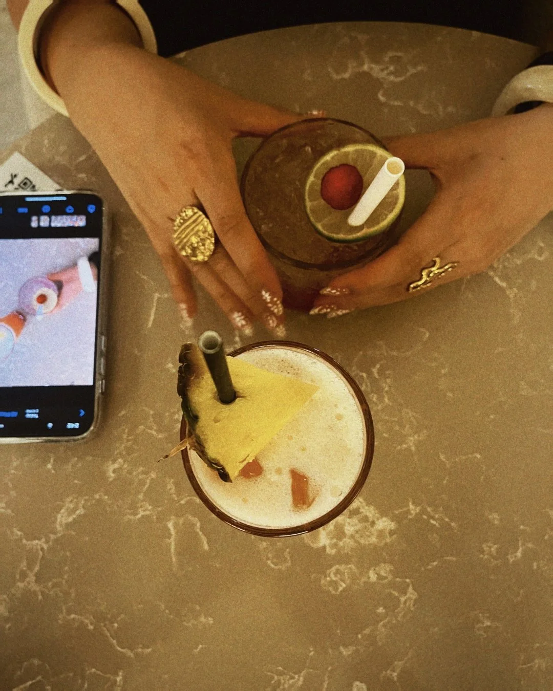 Deux mains tenant des verres de cocktail garnis de tranches d'ananas, avec une paille blanche dans chaque verre. Un téléphone sur la table affiche une photo de la même boisson. La table a un motif marbré beige ou crème.