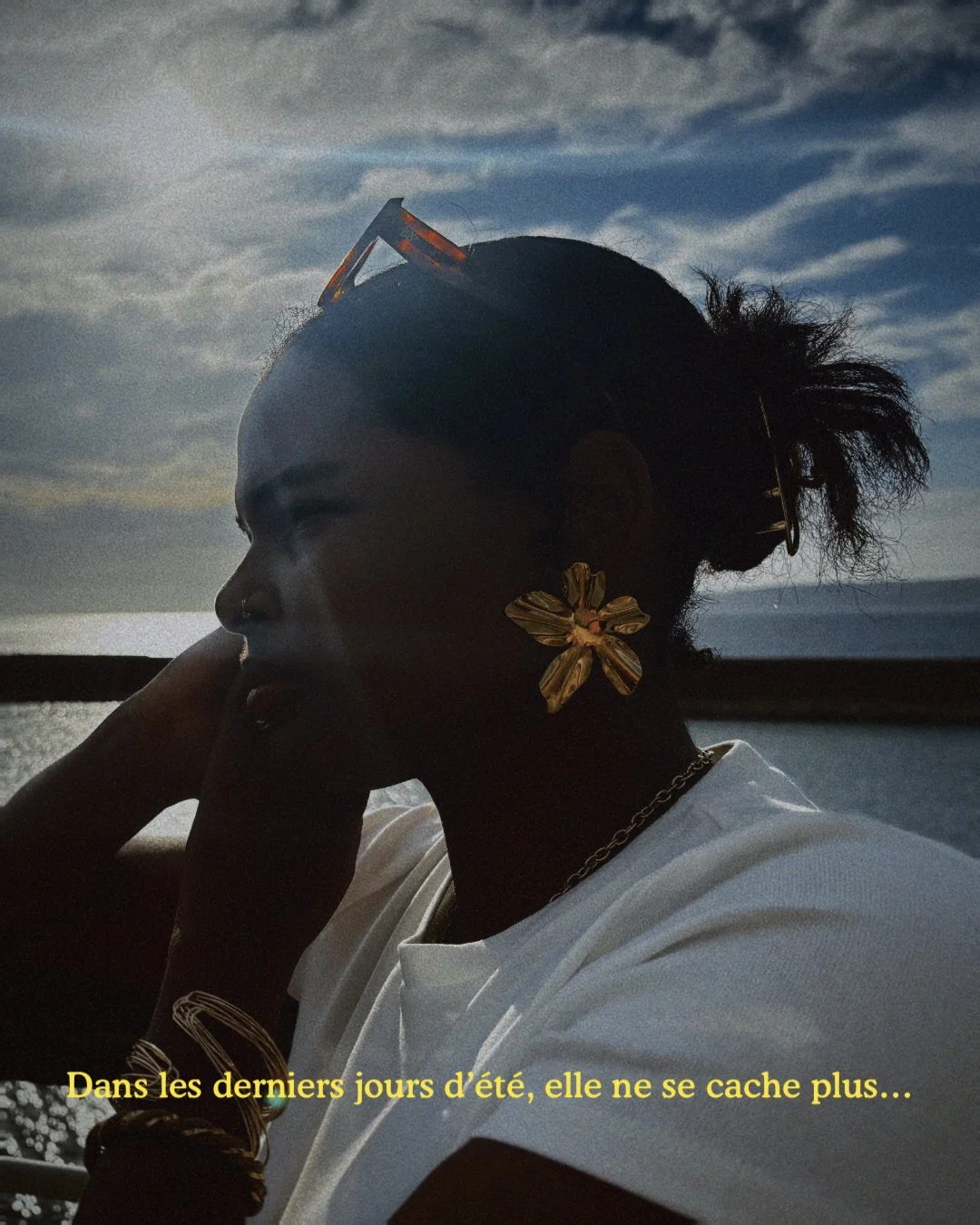 Portrait d'une femme avec une silhouette sombre, portant de grandes boucles d'oreilles en forme de fleur et des bracelets, regardant vers la gauche, avec un arrière-plan de ciel nuageux et de mer en fin d'été.