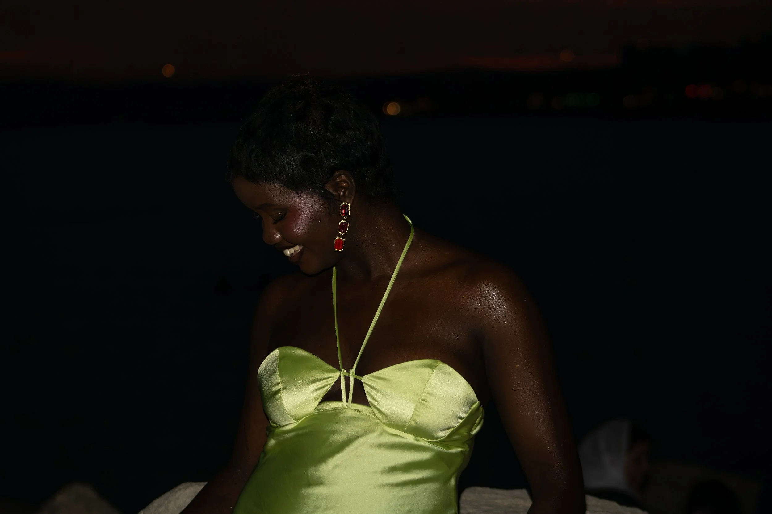 Une femme souriante porte une robe vert pastel avec un détail en forme de papillon et des boucles d'oreilles rouges, en pleine nuit.