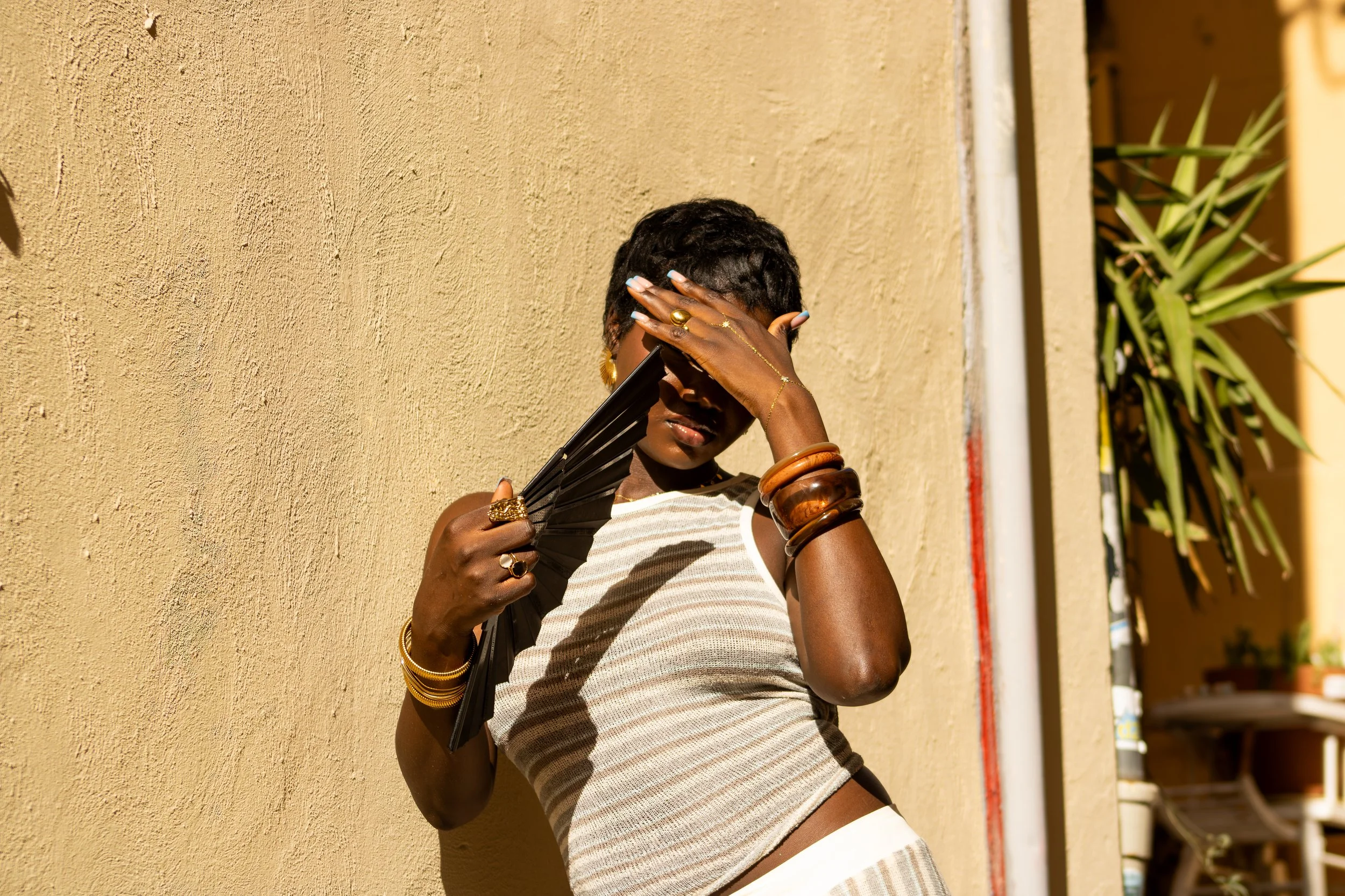 Femme noire portant des bijoux en acier inoxydable dorés, tenant un éventail noir, se tenant contre un mur beige au Panier à Marseille.