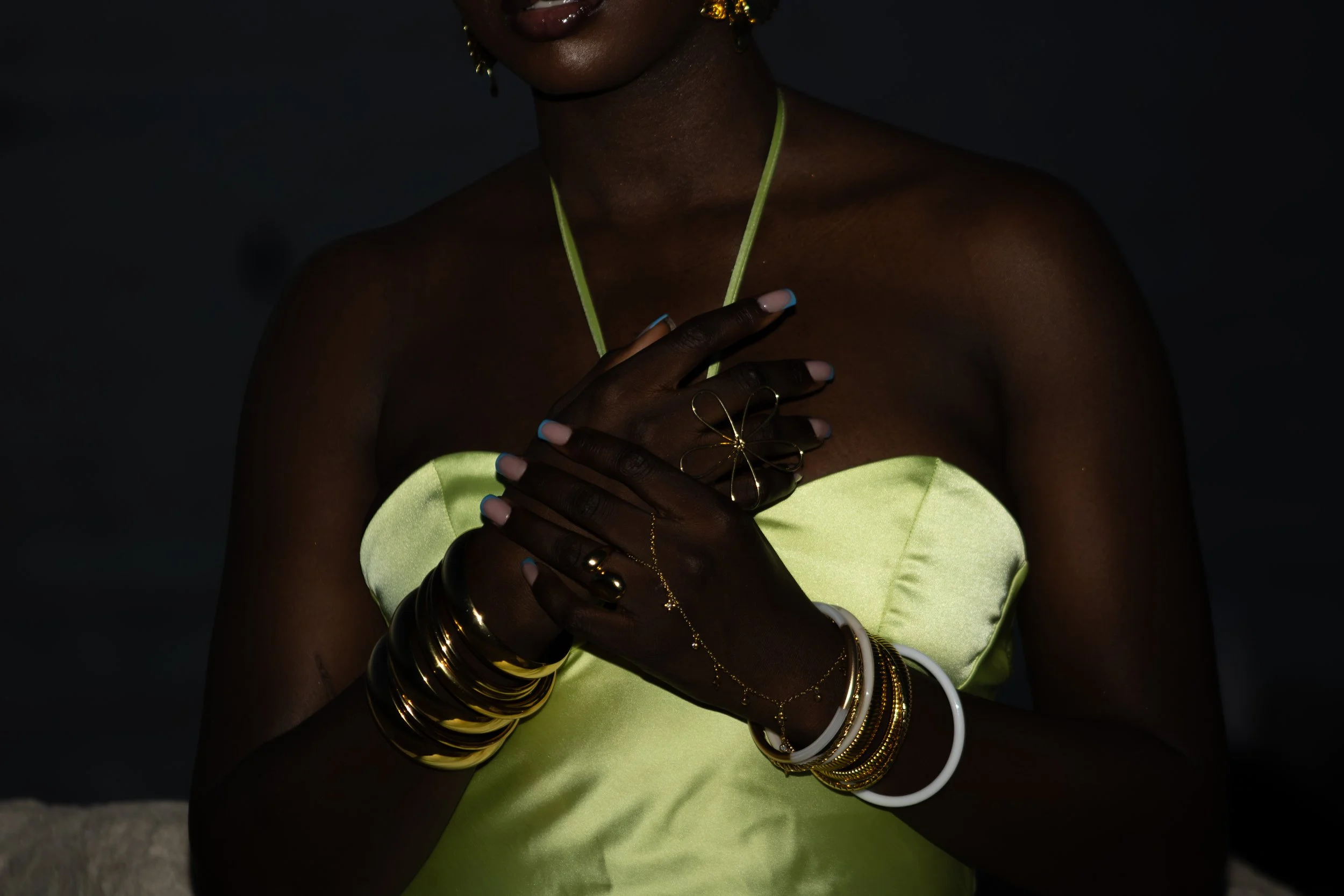 Une femme portant des bijoux en or et en argent, dont plusieurs bracelets et bagues, est vêtue d'une robe jaune fluo et tient la main sur sa poitrine.
