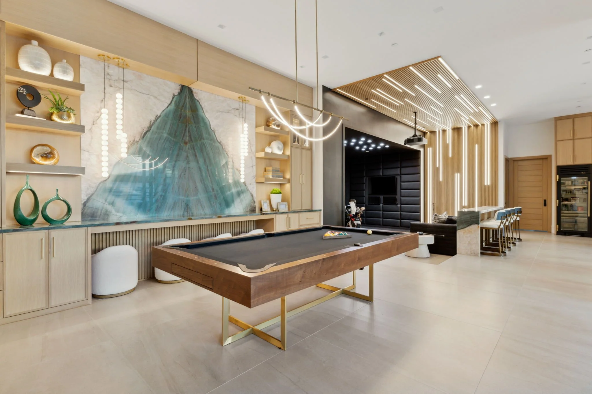 A modern recreational room with a pool table, decorative shelving, a large wall art, lighting fixtures, a TV, and a bar area with chairs.