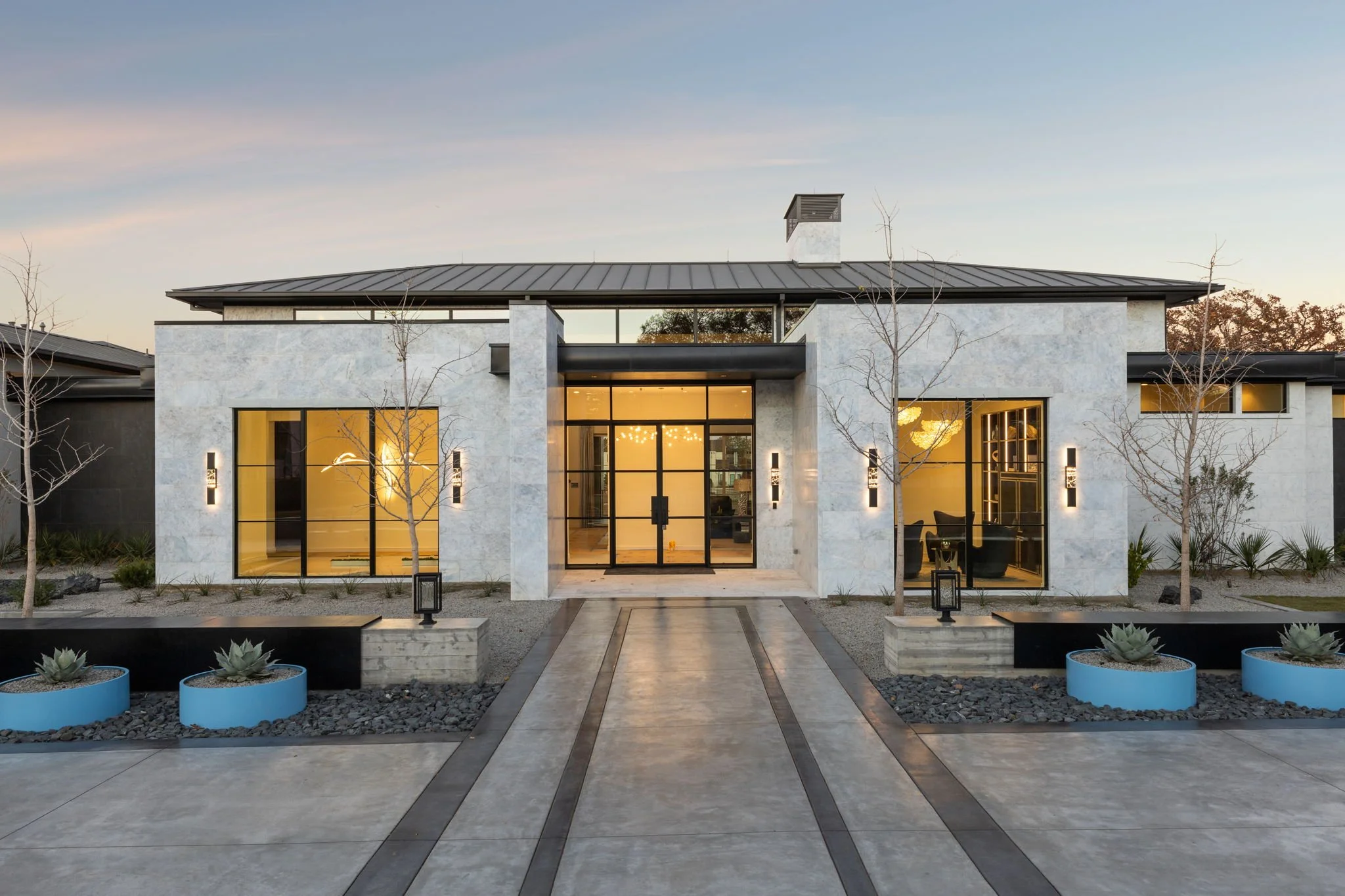 Modern house with white stone exterior, large black framed windows, and a metal roof, landscaped with small trees and planters, at dusk.