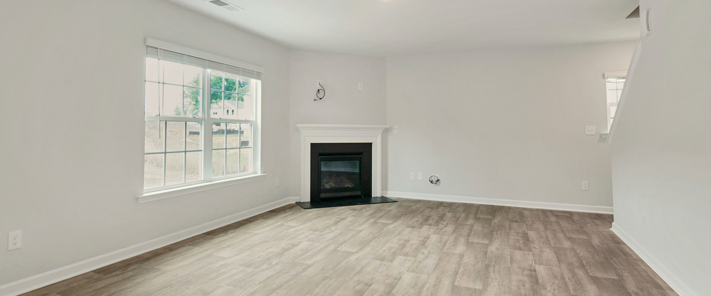 Empty living room with large window, white walls, gray hardwood floors, fireplace with white mantel, and some electrical outlets visible.