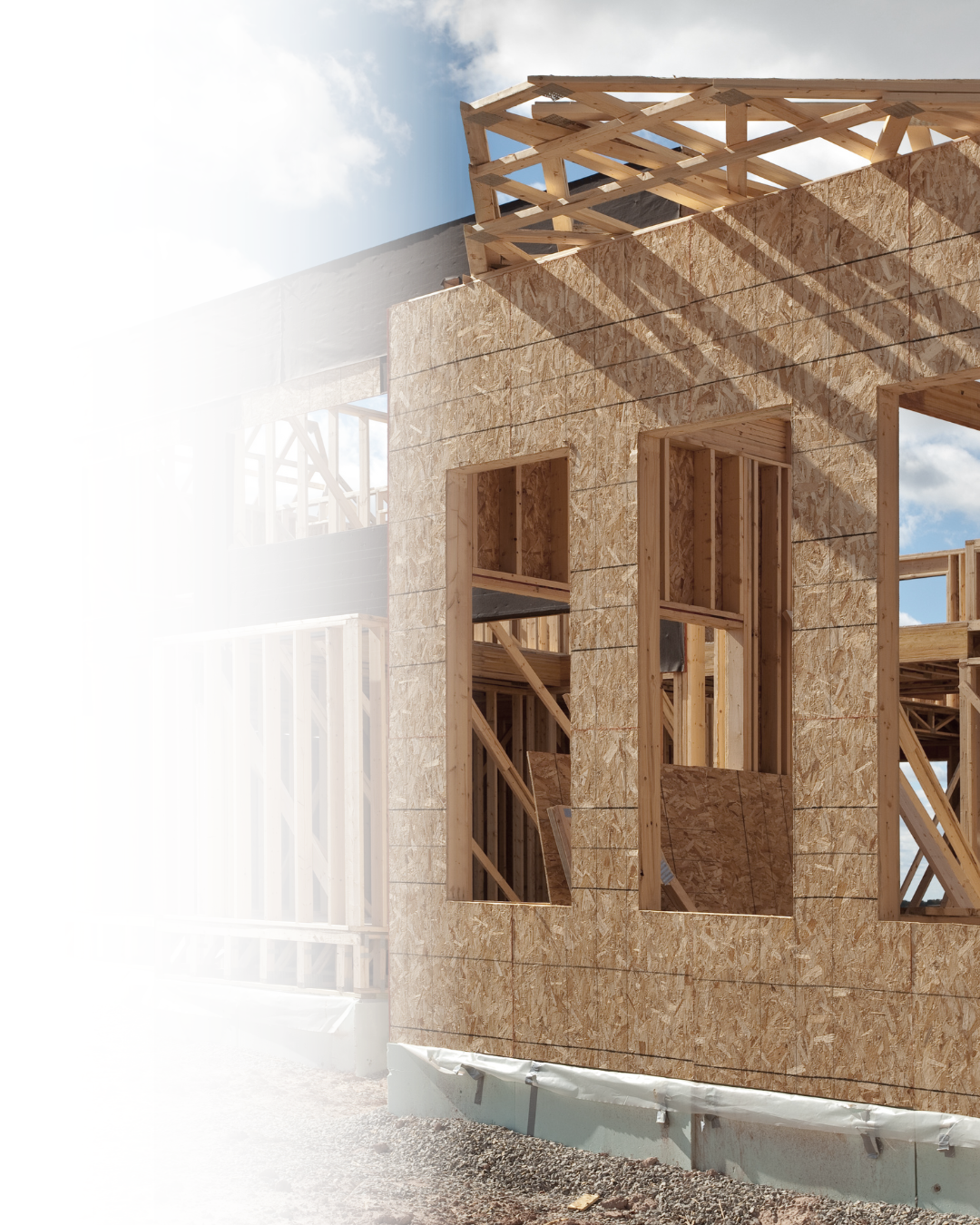 Wooden framing and exterior sheathing of a house under construction, with open window and door frames, and a partly built roof structure against a blue sky with clouds.