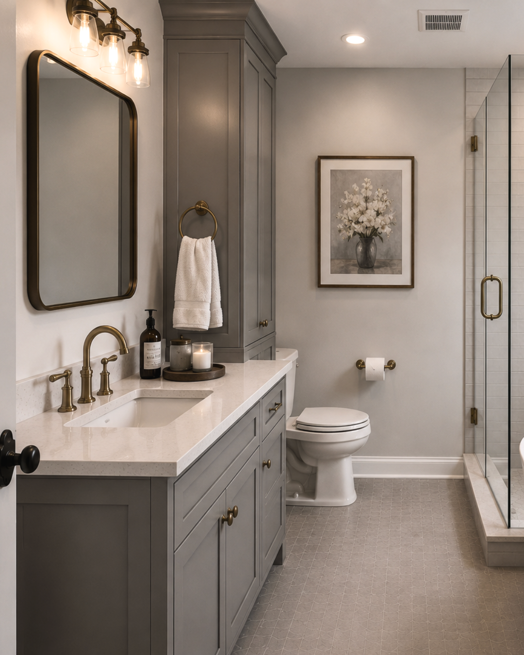 A modern bathroom with a gray vanity, gold fixtures, a white countertop, a mirror, a candle, a soap dispenser, a towel, a framed floral picture, a toilet, and a glass-enclosed shower.