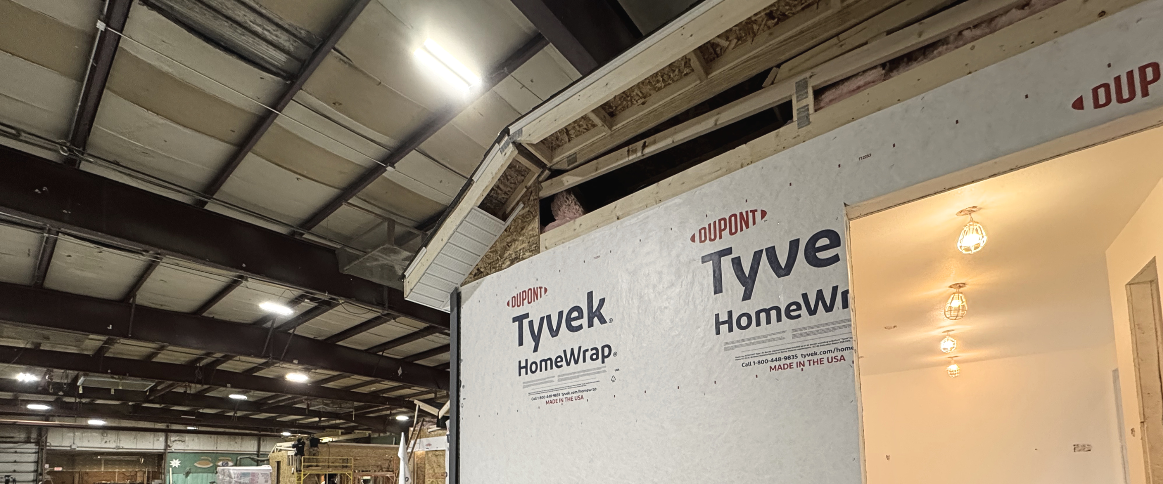 Interior of a building under construction with drywall showing branding from Tyvek and Dupont, exposed ceiling with metal beams, and a partially completed wall.