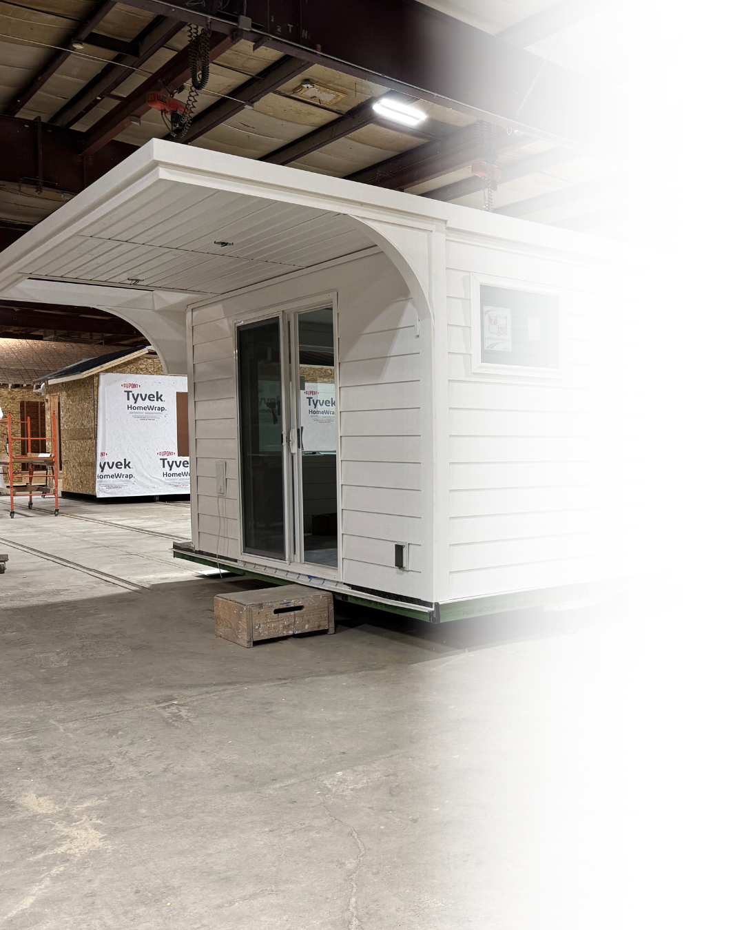 A white tiny house with a sliding glass door and small window, situated inside a warehouse or construction site with unfinished walls and exposed ceiling beams.