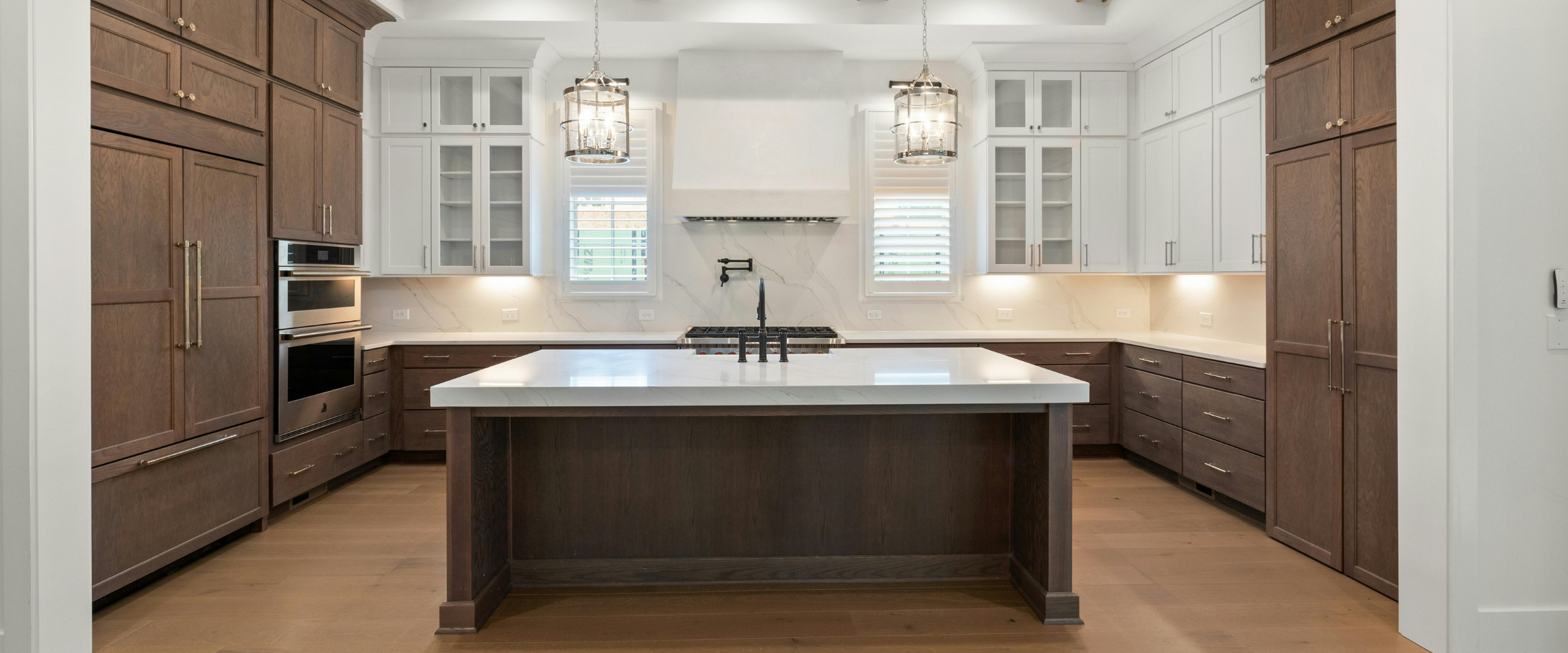 A modern kitchen with a large central island, wooden cabinets on the left and right, white upper cabinets with glass doors, and a marble backsplash. There are two windows with shutters, pendant lights hanging above the island, and stainless steel appliances including double ovens and a stove. The floor is wood, and the overall style is clean and contemporary.