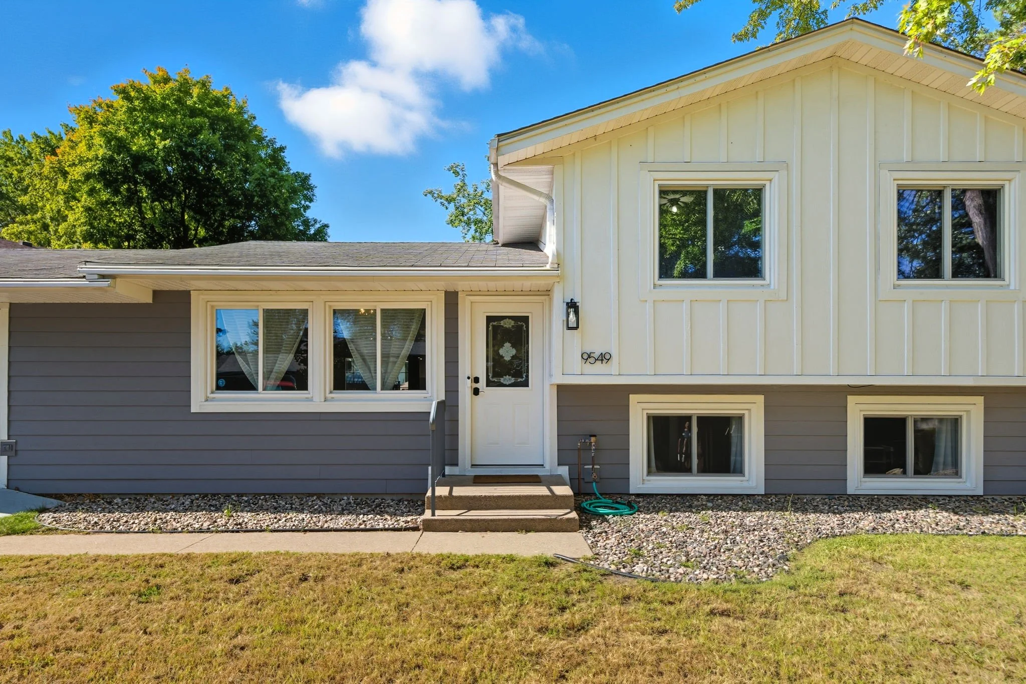 Front exterior of a two-story house with a green lawn, a concrete walkway, and a house painted in white and dark gray siding. The house has three windows on the upper floor, a front door with a small black railing, and a window below the porch. 