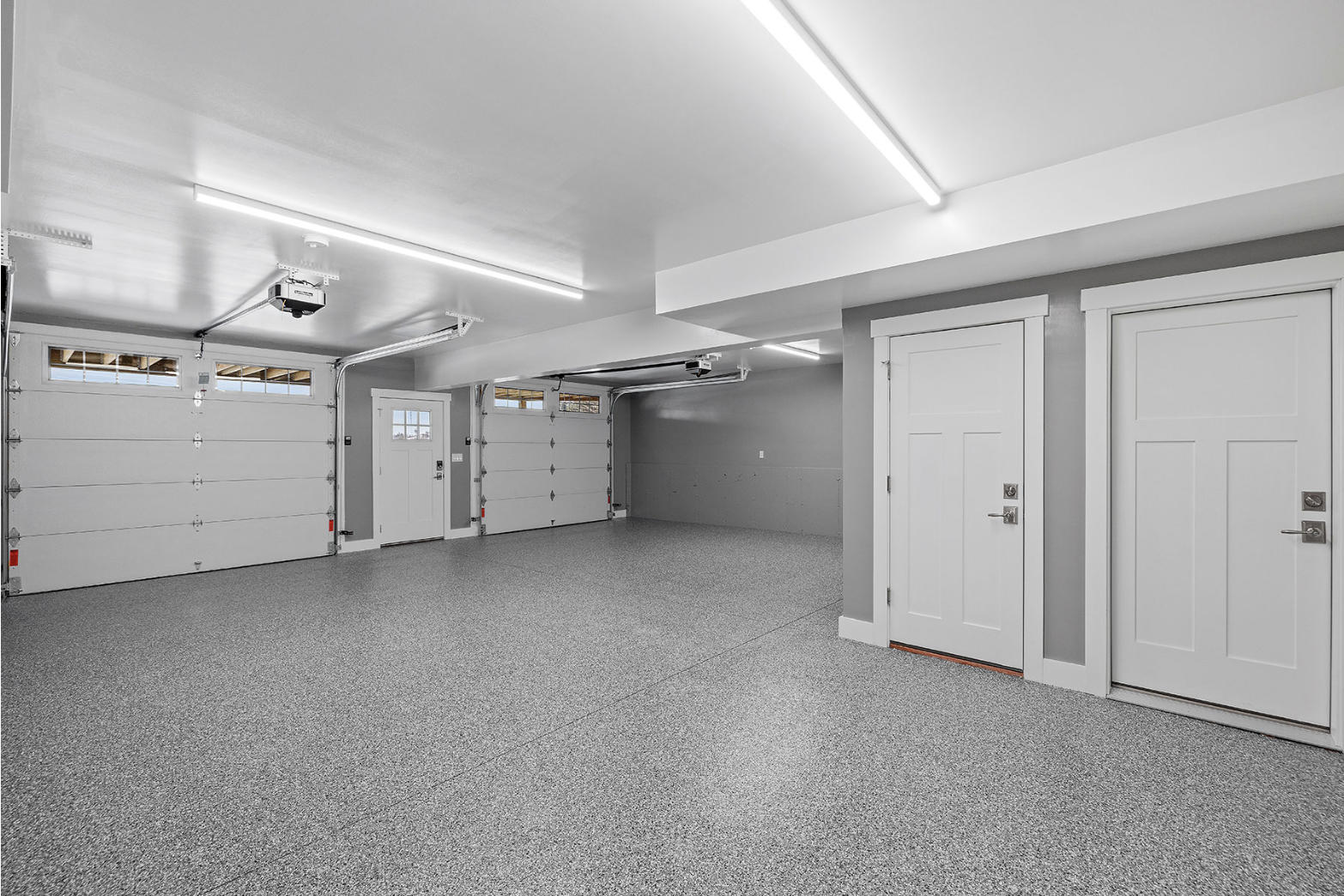 Empty two-car garage with overhead doors, white walls, and gray speckled epoxy flooring.
