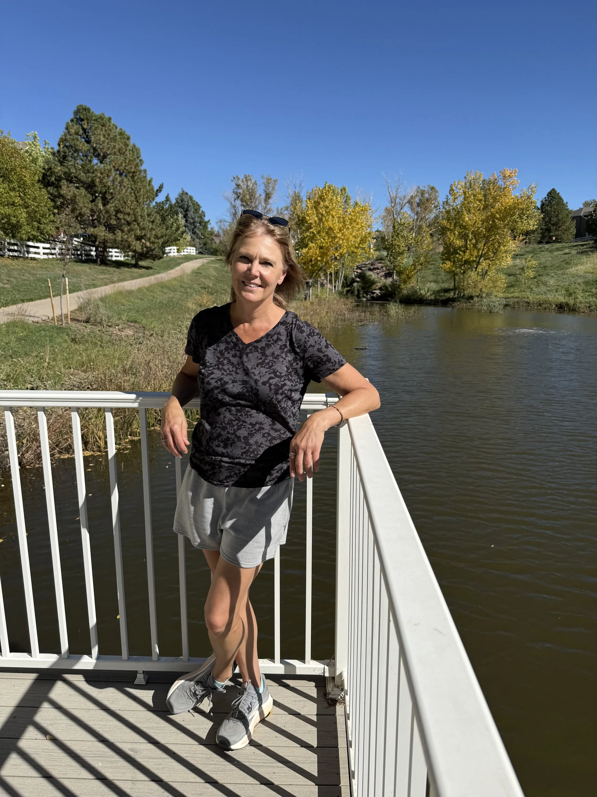 Stacey, pain recovery coach at Your Path Out Of Pain, smiling on a sunny bridge over a pond with fall trees behind her.