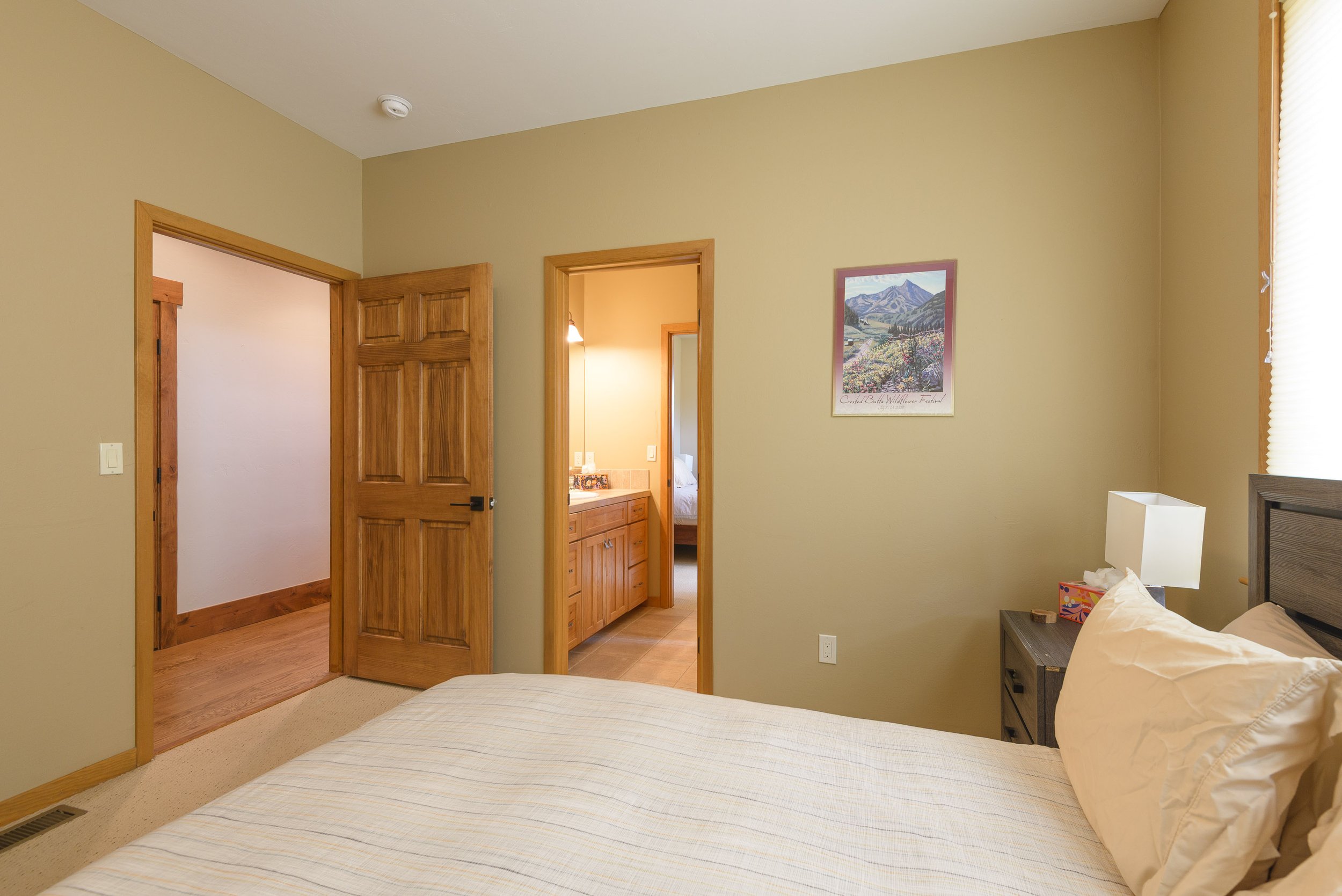A bedroom with beige walls, a wooden door leading to another room, a small dresser with a lamp, lampshade, tissue box, and a bed with pillows.