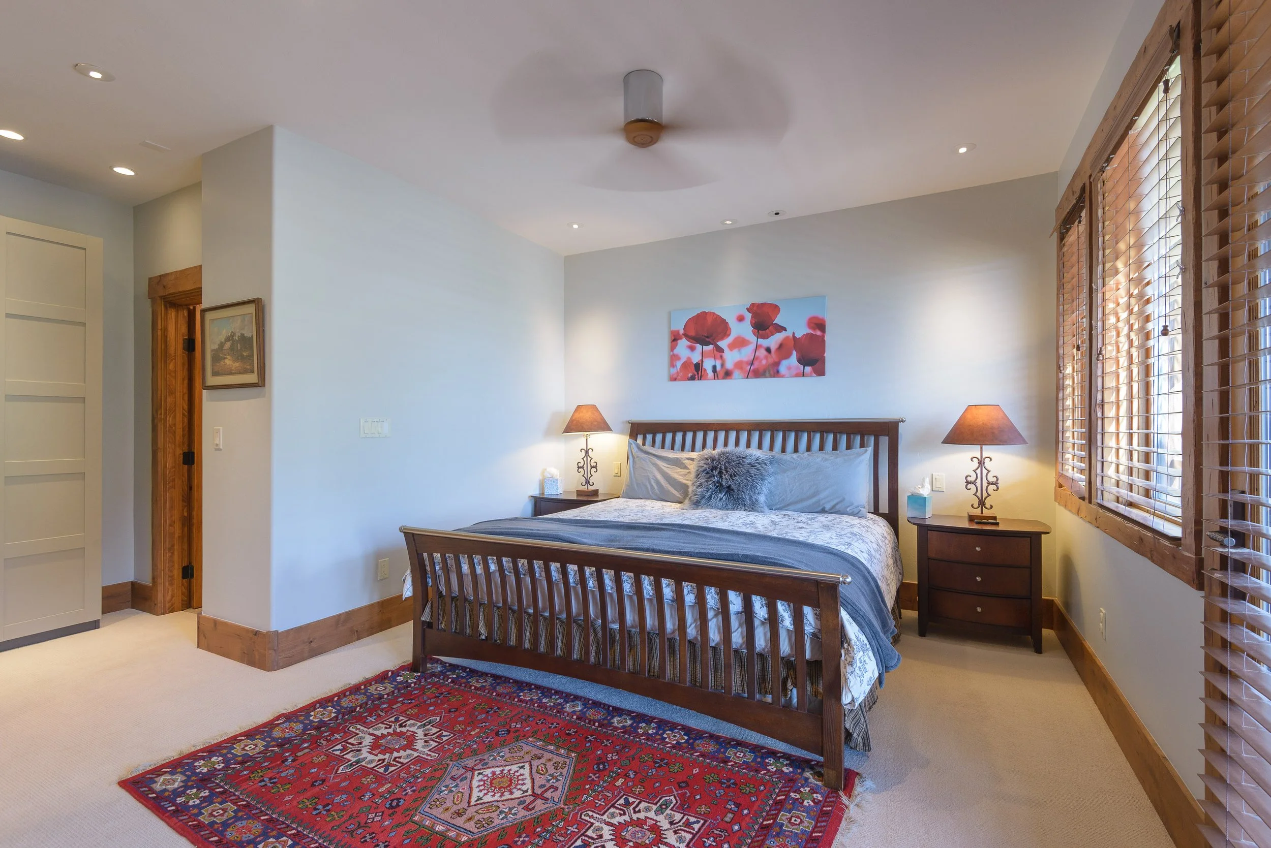 A bedroom with a wooden bed, two bedside tables with lamps, a colorful rug, and a painting of flowers on the wall.