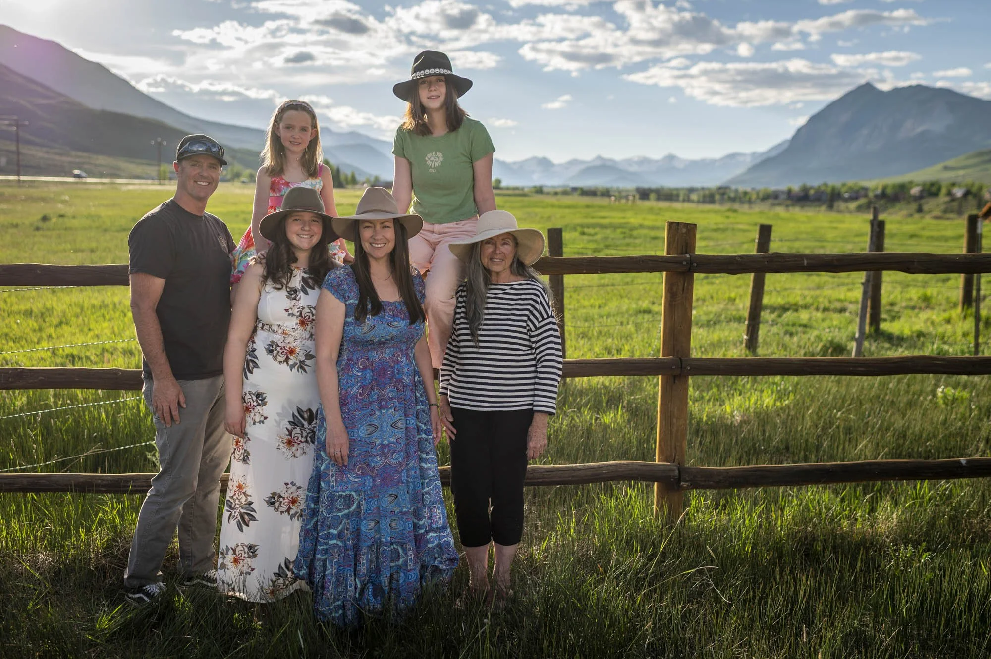Family of seven outdoors in a grassy field with mountains in the background, wearing summer clothes and hats, smiling for the photo.