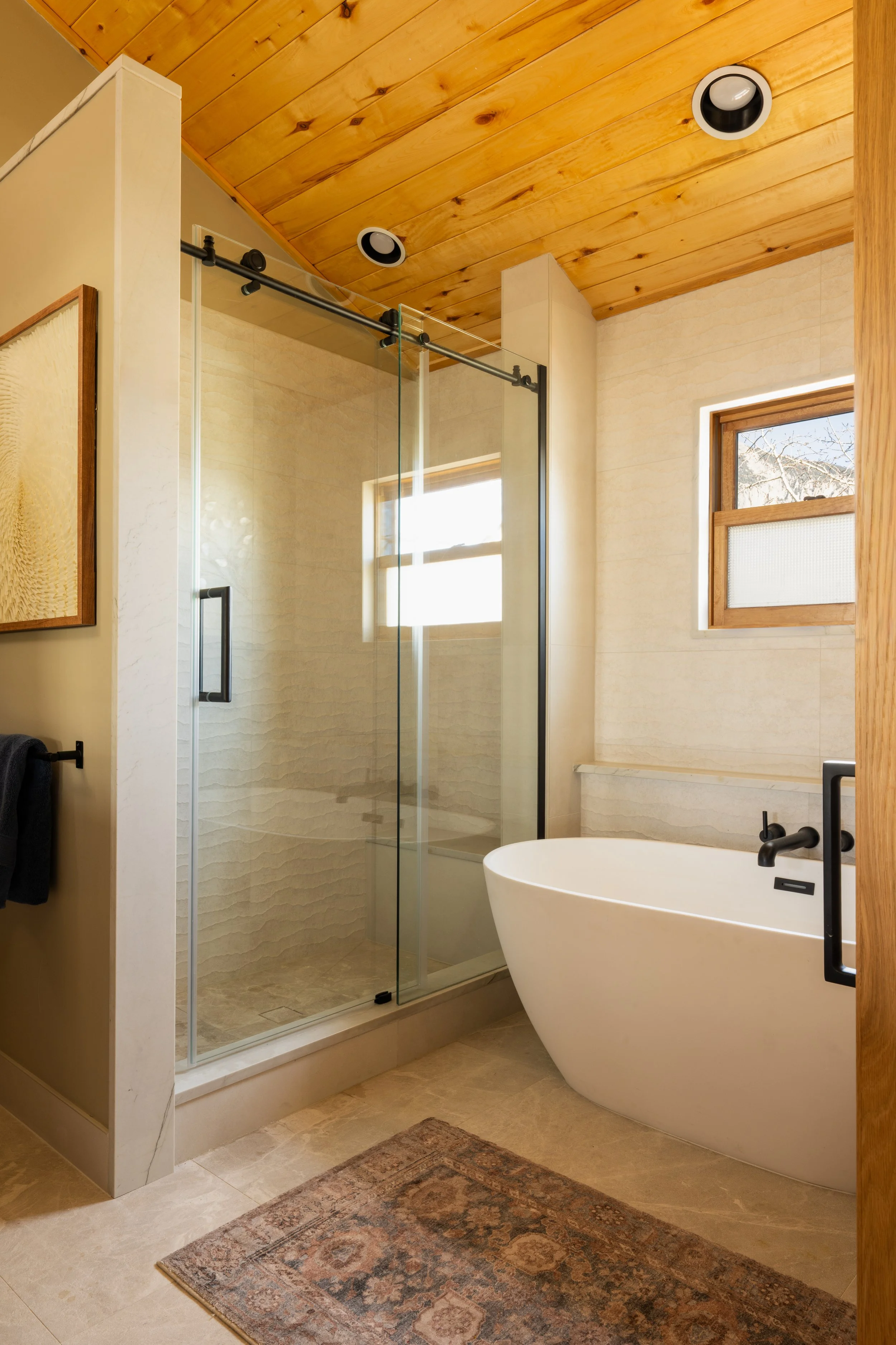 A modern bathroom with a wooden ceiling, a glass shower enclosure, a white oval bathtub, and small windows with natural light.