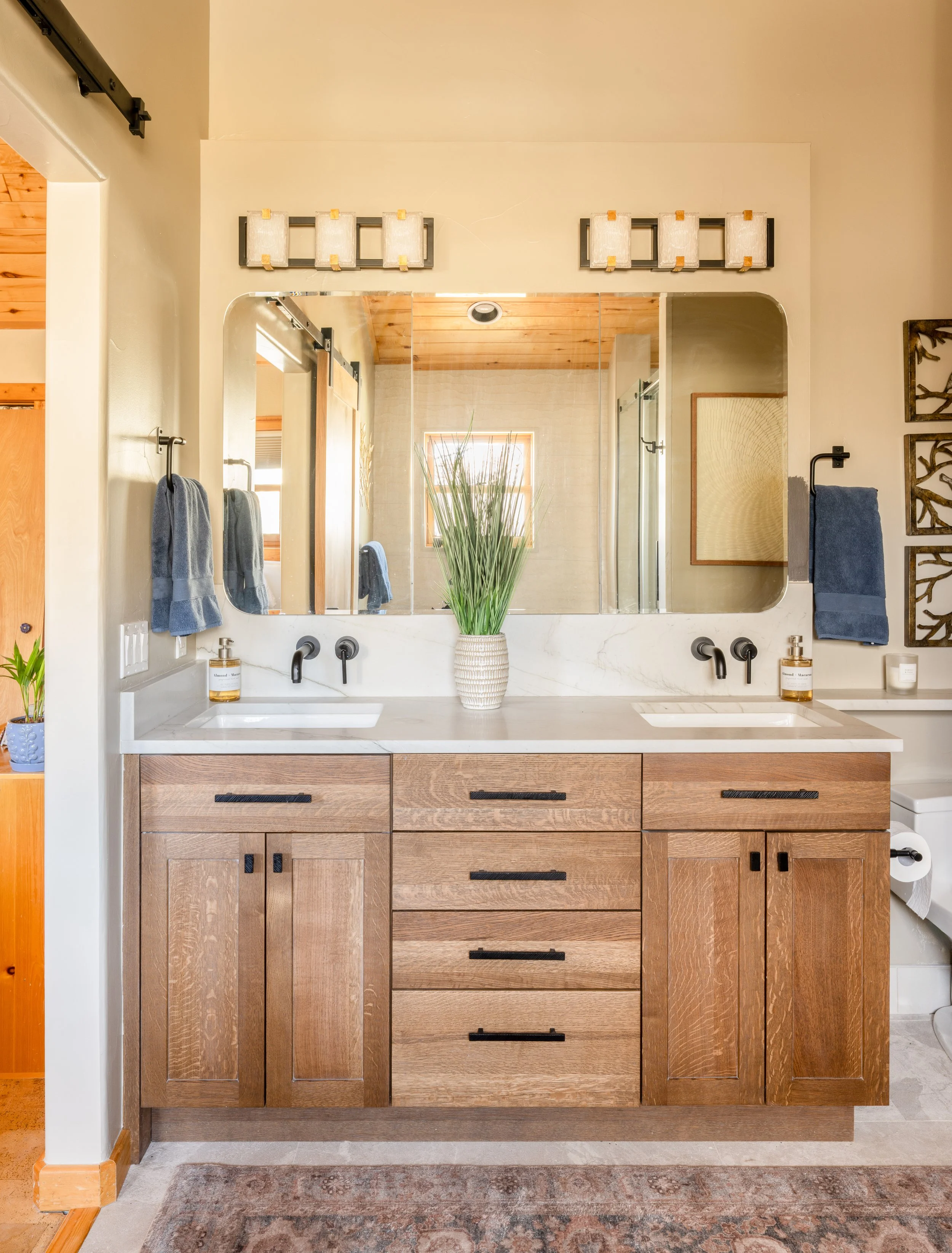 Bathroom vanity with double sinks, large mirror, wooden cabinets, a plant in a decorative vase, and towel hooks with blue towels.