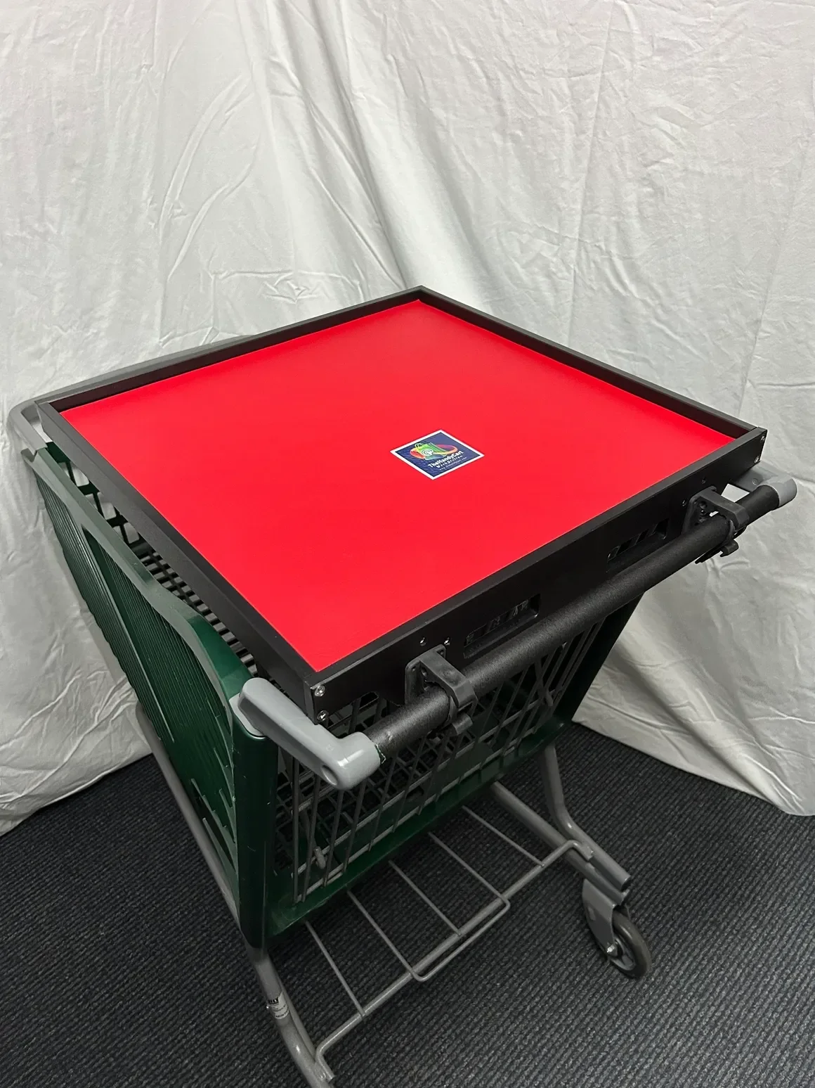 A red square tabletop with a small sticker in the center, mounted on a black frame, placed on top of a green shopping cart in front of a white fabric background.