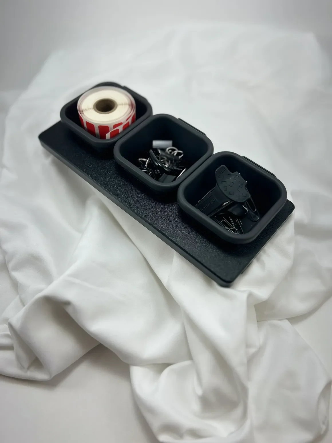 Three black square containers holding various items are placed on a black tray, with white fabric underneath. The first container has a roll of red and white striped tape, the second contains black and metallic small hardware pieces, and the third ha