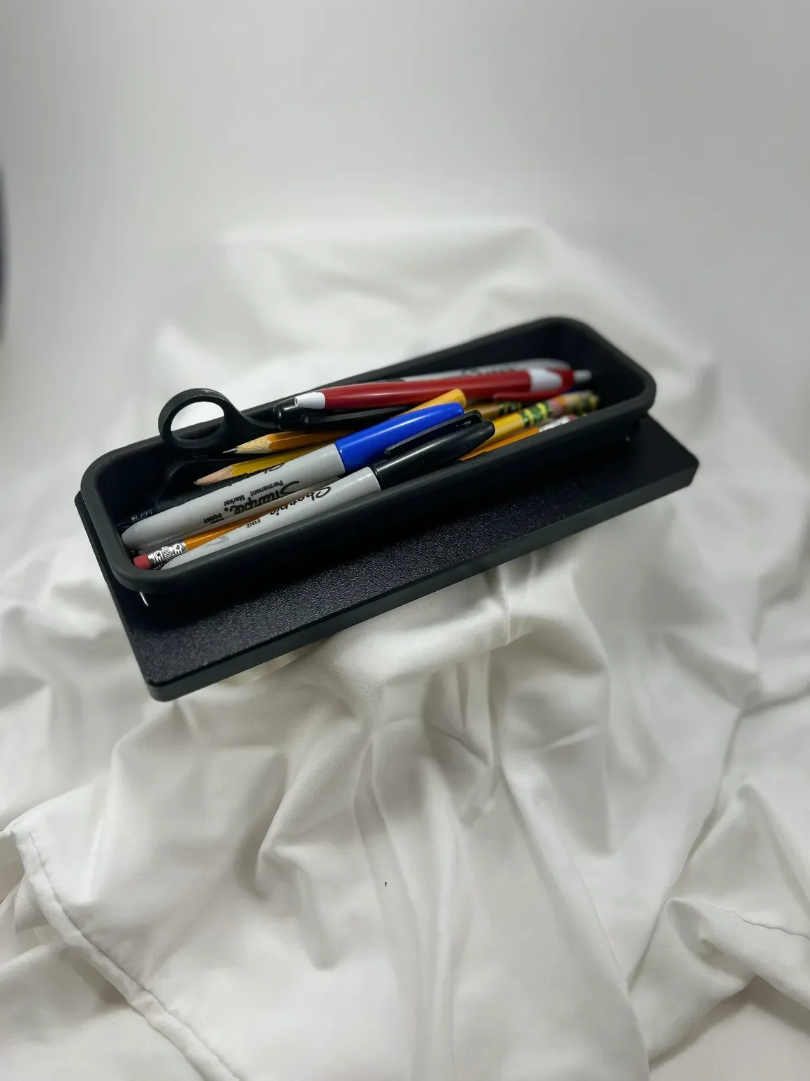 A black tray filled with various pens, pencils, and an eraser, resting on crumpled white fabric.