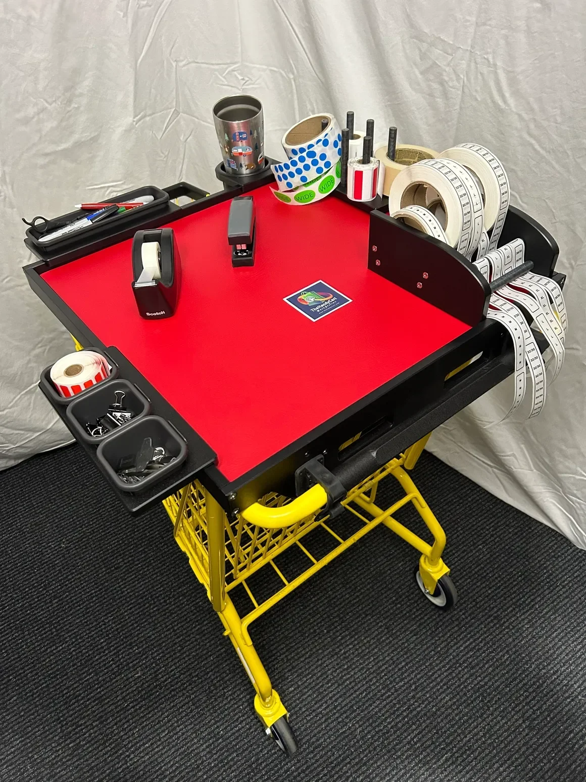 A red tabletop shopping cart with various packing supplies such as tape, scissors, pens, and measuring tape, set against a white backdrop.