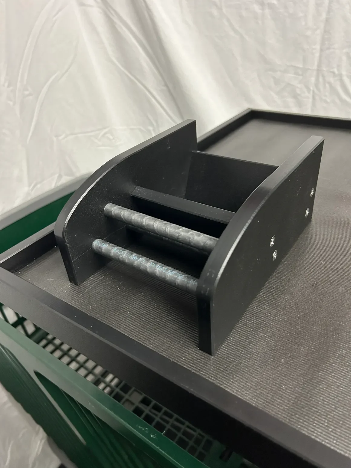 A black, rectangular metallic device with two parallel cylindrical rollers and a small side compartment, placed on a dark textured surface in front of a white backdrop.